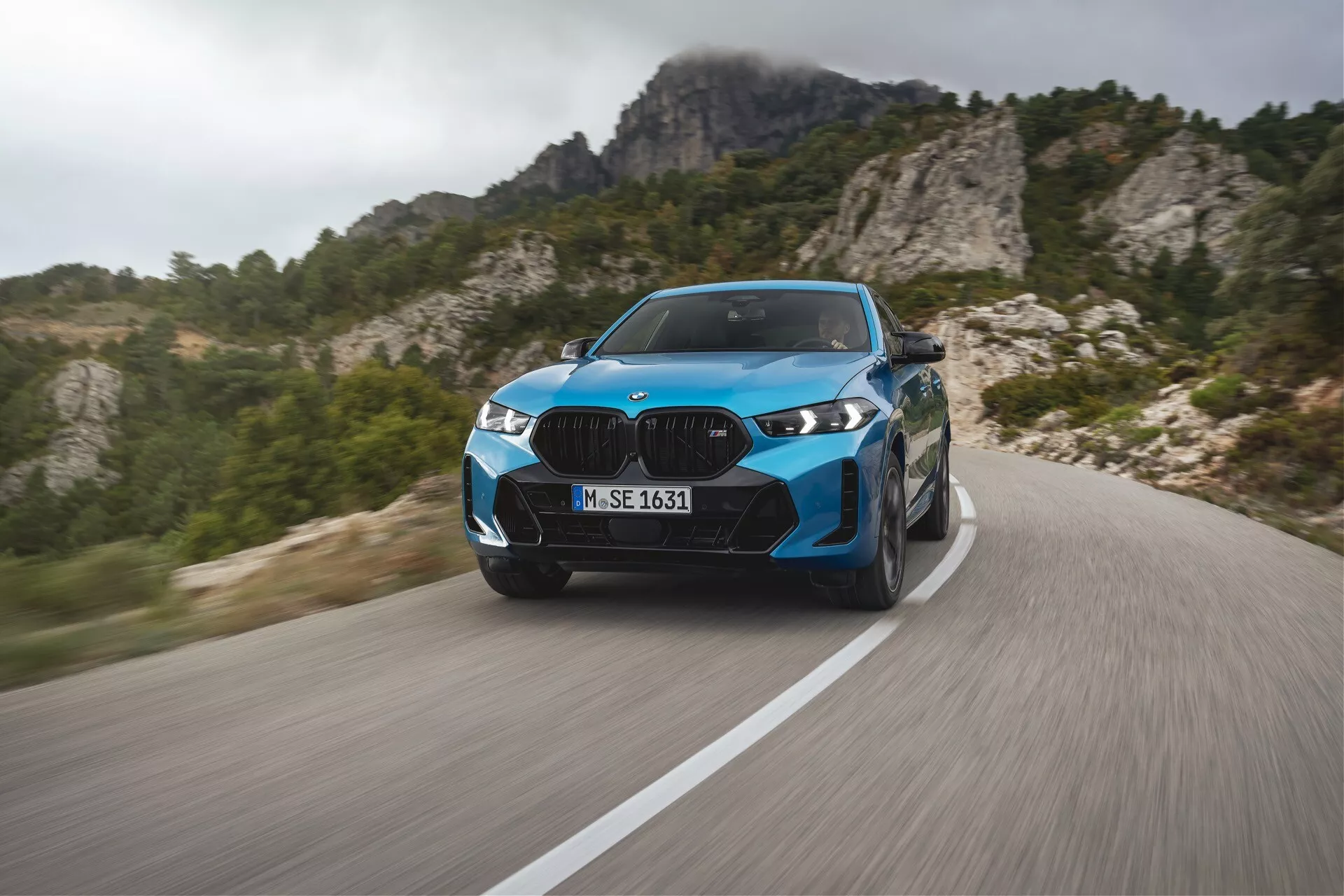 BMW X6 2023 (G06-F96-facelift) en bleu vibrant, illustrant une performance de luxe moderne sur une route de montagne sinueuse avec flou de mouvement dynamique, phares LED avancés, éléments de design élégants et roues sportives, sur un fond de montagnes escarpées.