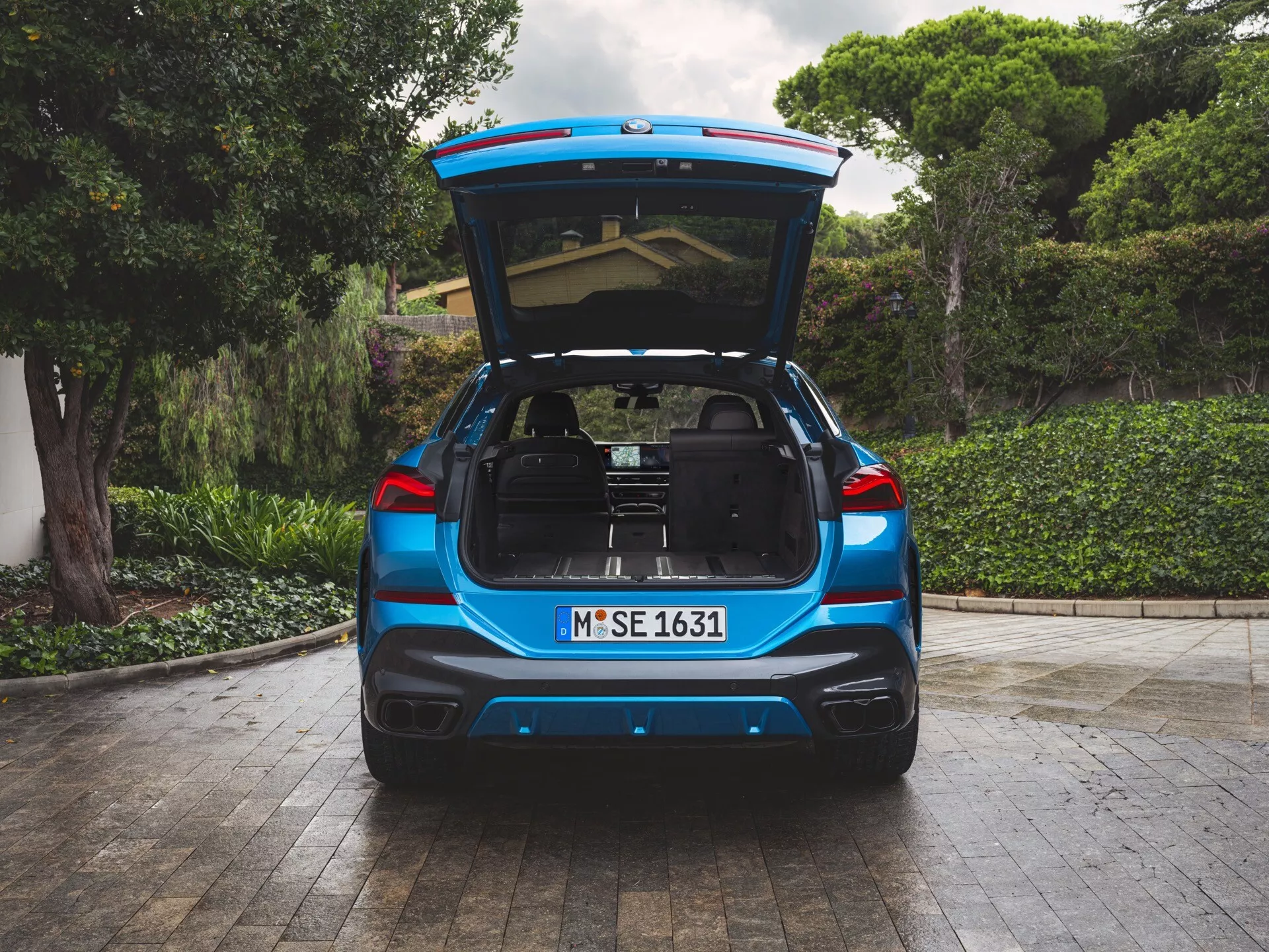 Vue arrière du BMW X6 2023 (G06-F96-Facelift) en peinture métallique bleue, coffre ouvert révélant un espace de chargement spacieux, garé sur un pavage en pierre avec de la verdure et une partie de maison en arrière-plan, avec feux arrière LED, échappements jumeaux, et possibles technologies d'assistance au stationnement