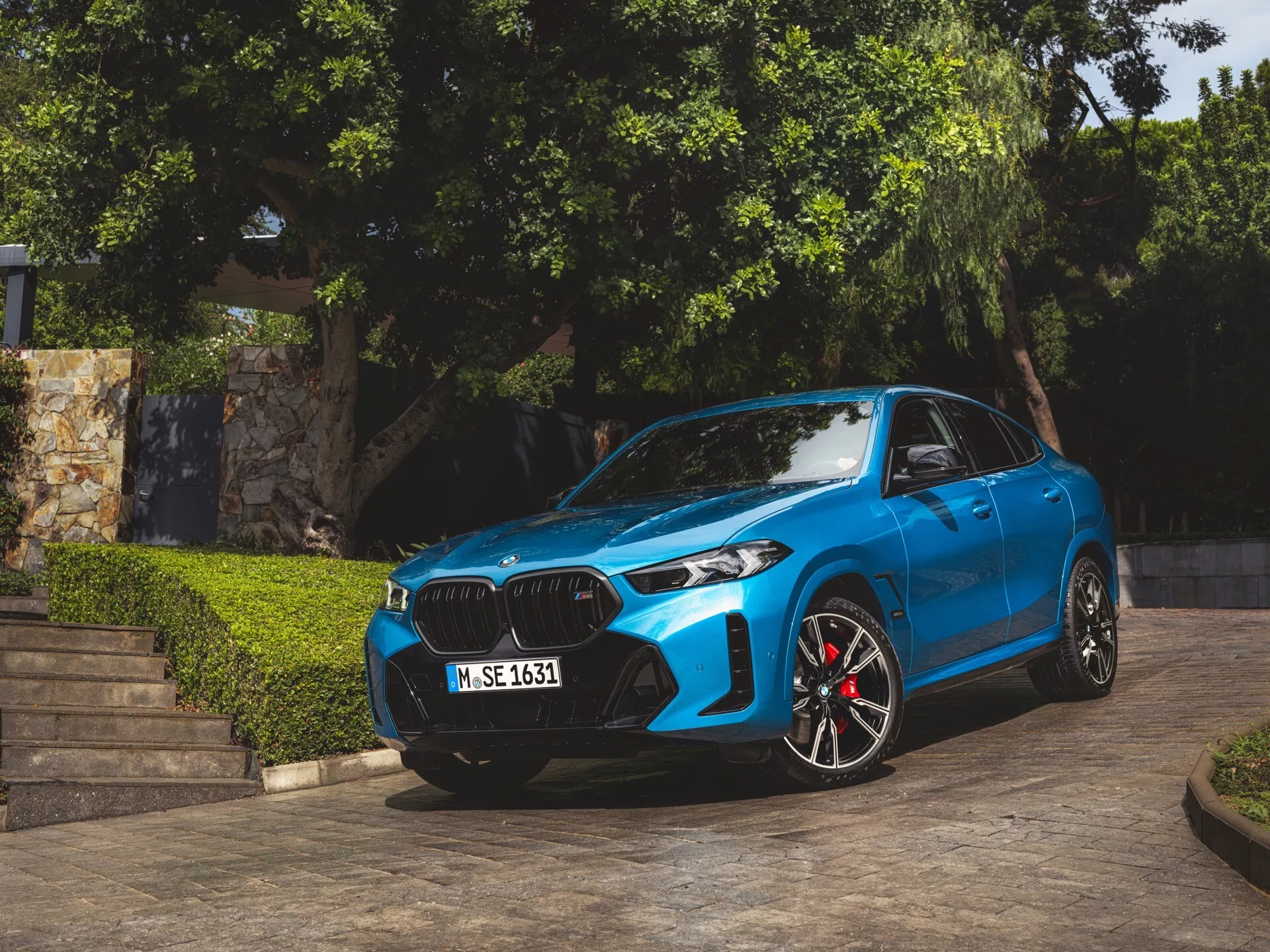 BMW X6 2023 (G06-F96-Facelift) en bleu, garée en angle montrant sa façade avant, grille en forme de rein, phares LED ou laser, roues sportives en alliage avec étriers de frein rouges, lignes contourées sur le capot et rétroviseurs latéraux aérodynamiques avec clignotants intégrés, capturée à la lumière naturelle du jour avec de la végétation et des marches architecturales en arrière-plan