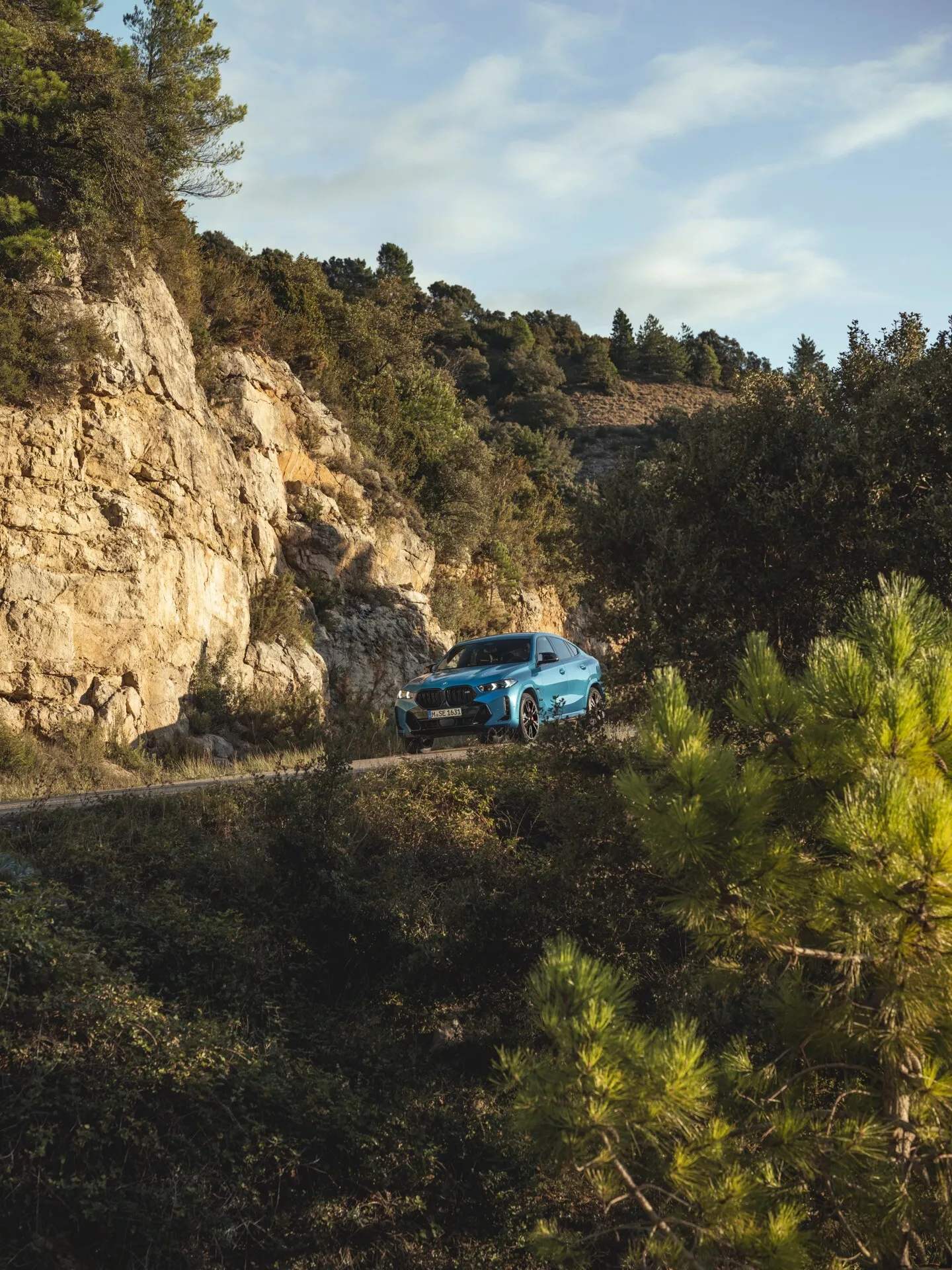 BMW X6 2023 (restylage G06-F96) vue de trois quarts avant sur une route de montagne sinueuse entourée de forêt, démontrant capacité tout-terrain et design moderne