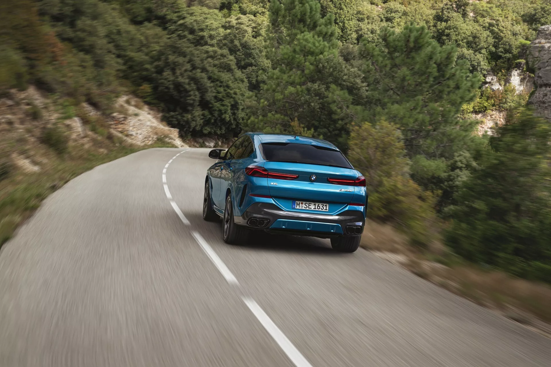 Vue arrière dynamique d'une BMW X6 2023 (G06-F96-Facelift) bleue, présentant un design sportif et des feux arrière modernes sur une route sinueuse au milieu d'un paysage naturel