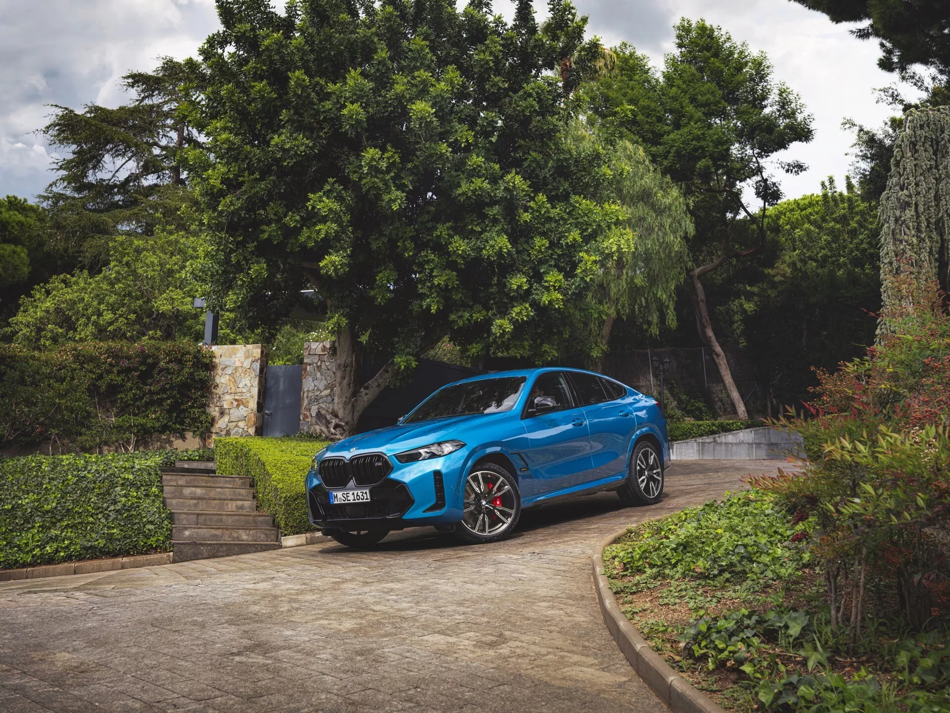 BMW X6 2023 (G06-F96-Restylage) en bleu, vue de trois quarts avant, garée sur une allée entourée de verdure, montrant pare-chocs avant, grille, capot, pare-brise, profil côté conducteur, phares LED, jantes sportives avec étriers de frein rouges, et design aérodynamique