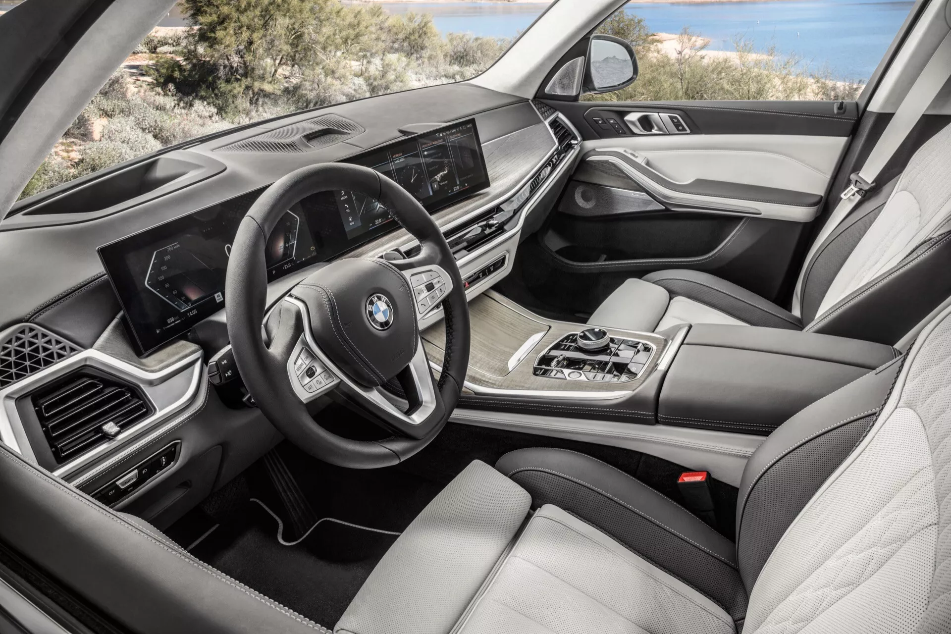 Vue intérieure d'une BMW X7 2022 (G07-Facelift) du côté passager mettant en vedette le tableau de bord avec écran d'infodivertissement, volant multifonction, levier de vitesse automatique, commande multimédia iDrive, contrôle de climatisation bi-zone, sièges en cuir de luxe, ornements en bois et métal et cluster d'instruments digital.