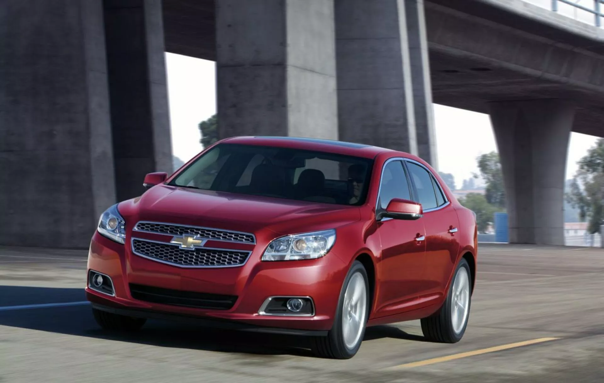 Vue dynamique d'une berline Chevrolet Malibu 2012 rouge en mouvement sur un pont, présentant un design frontal avec grille chromée, phares élégants, silhouette sportive et jantes en alliage.