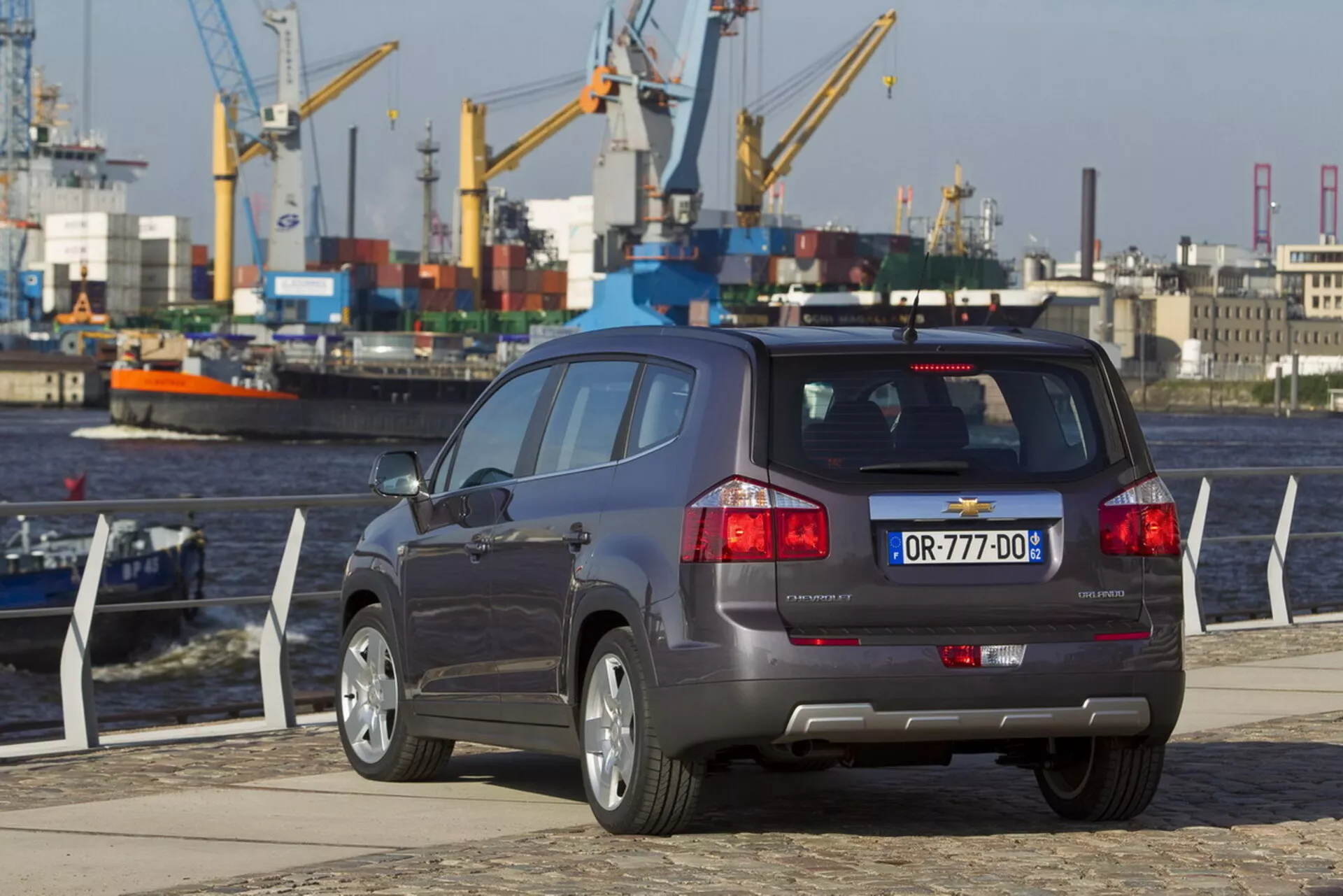Vue arrière trois quarts de la Chevrolet Orlando 2011 sur un quai en bord de l'eau, mettant en avant sa carrosserie de SUV compact, configuration à cinq portes, système d'échappement double, capteurs d'aide au stationnement sur le pare-chocs, et jantes en alliage à multiples branches