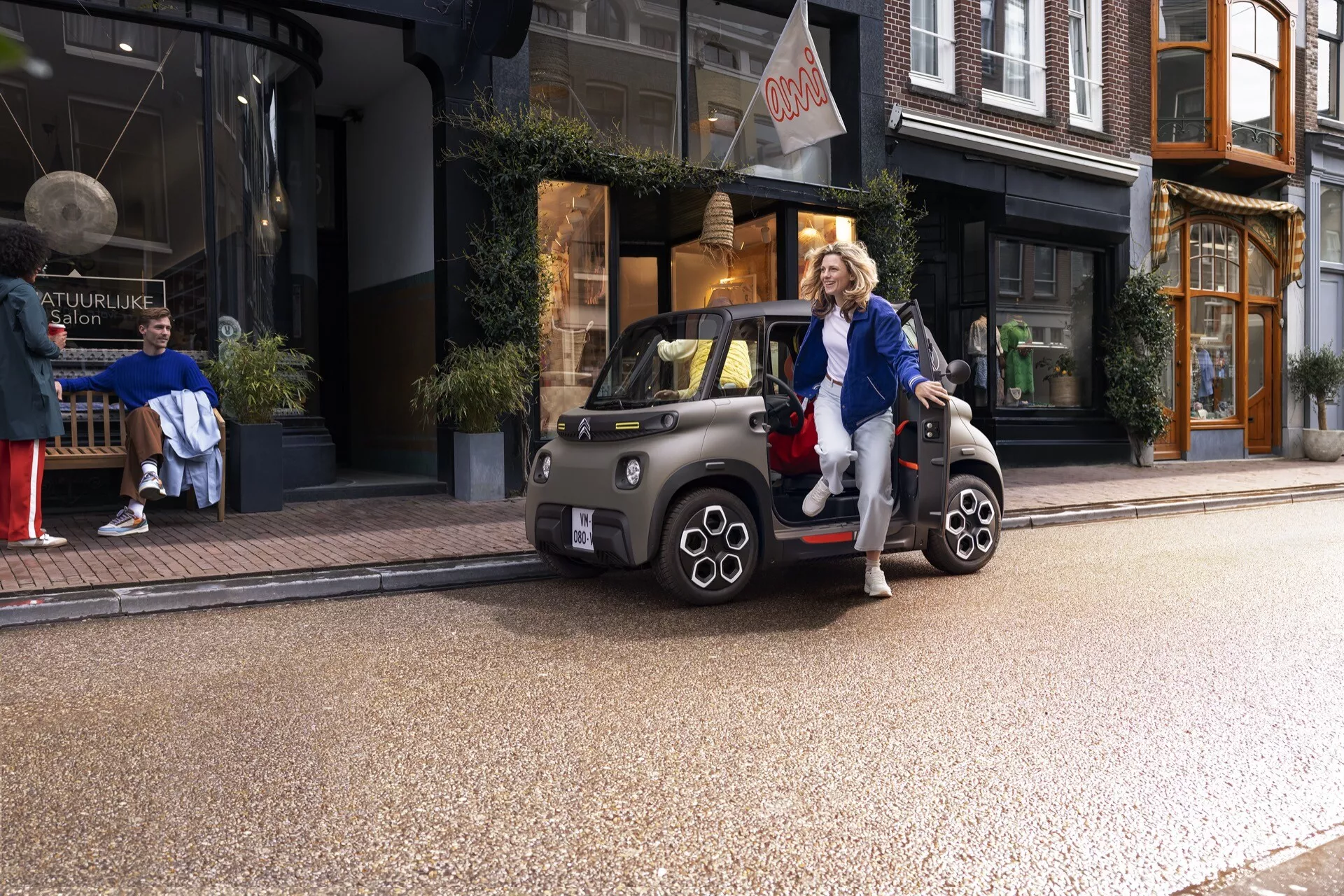 Le Citroen Ami 2024 est représenté dans une vue de trois quarts avant, montrant son design moderne et original avec des phares ronds et une grille ondulée, dans un environnement urbain avec des boutiques en arrière-plan. La voiture est un cabriolet avec le toit rétracté et sans portes, permettant une vision claire des petites roues multibranches et des jantes bicolores. Une personne habillée de manière décontractée à côté du véhicule ajoute un élément de style de vie, soulignant son image éco-responsable et urbaine.