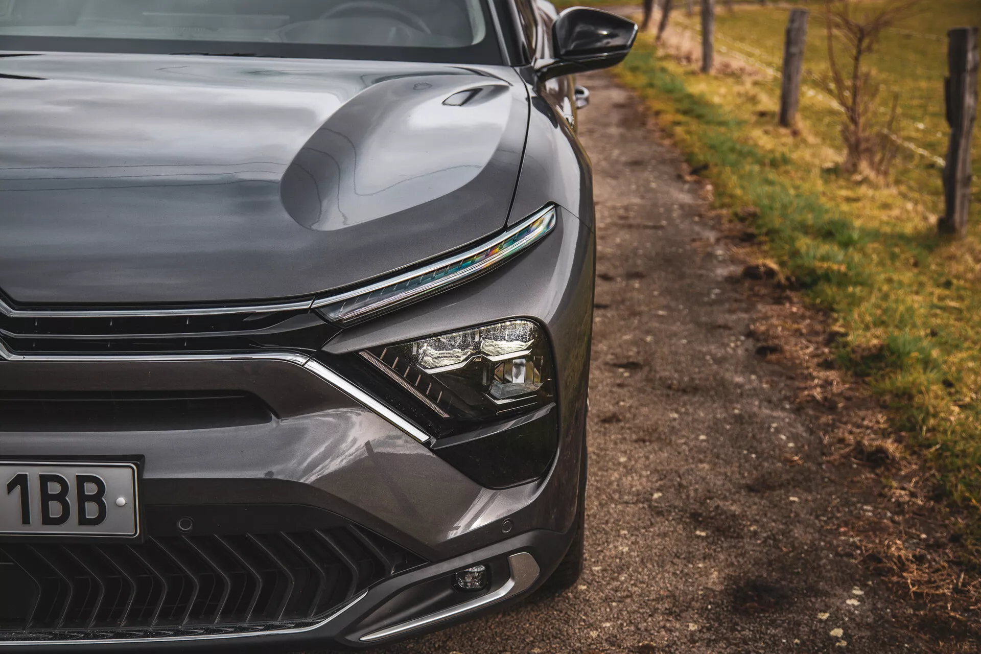 Gros plan sur la Citroen C5-X 2022 montrant le design élégant du côté avant gauche, y compris le capot profilé, la grille prononcée avec des lignes horizontales, le système de phare LED avancé et le pare-chocs avant façonné de manière aérodynamique avec des entrées d'air, sur un arrière-plan naturel flou en bordure de route.