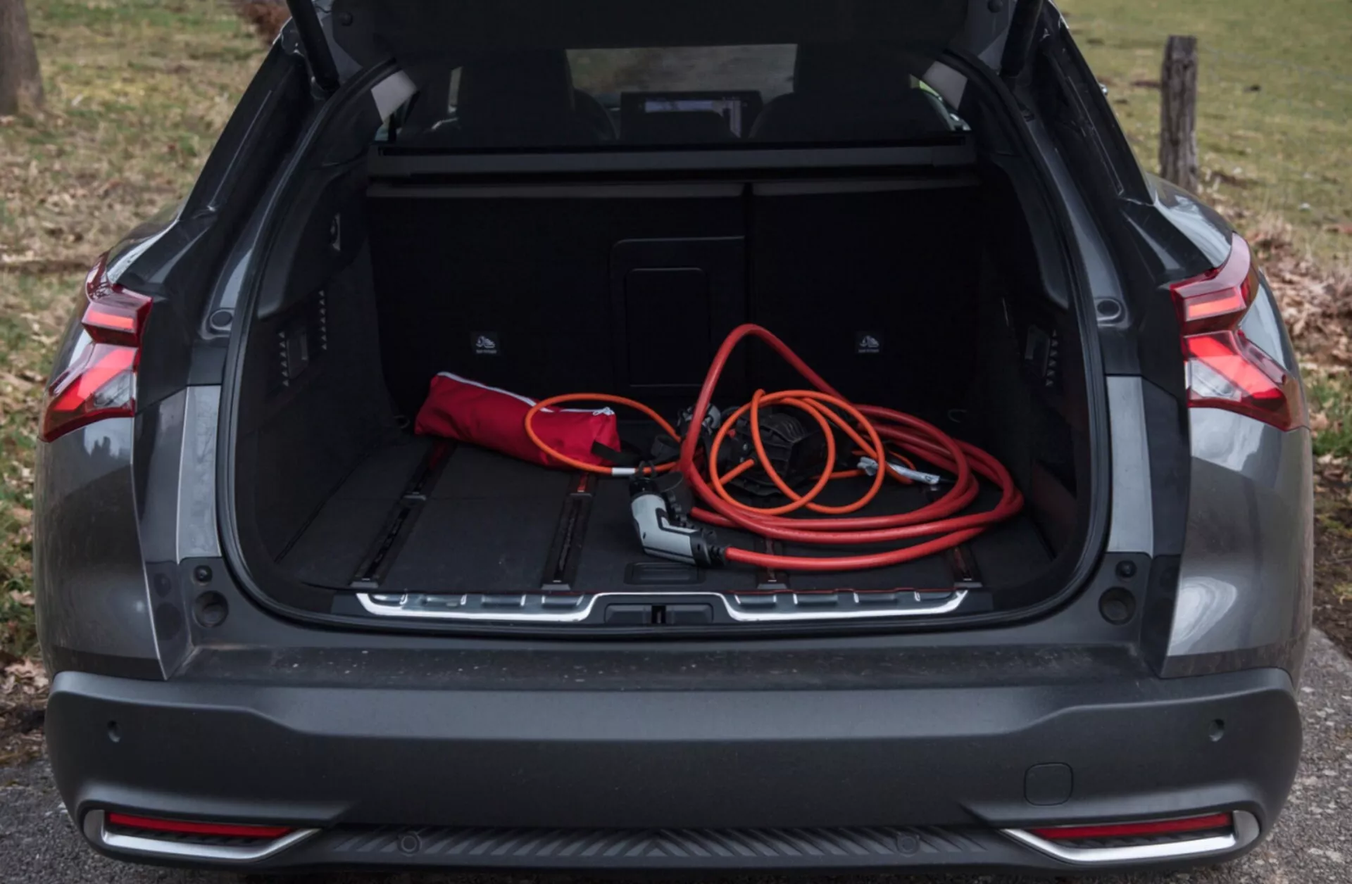 Vue de l'espace du coffre arrière de la Citroën C5-X 2022 avec compartiment ouvert montrant un espace de chargement spacieux, sac rouge et noir, cordon de rallonge orange, recoins de rangement supplémentaires, garniture de protection, et capteurs de stationnement, sous un éclairage naturel en extérieur