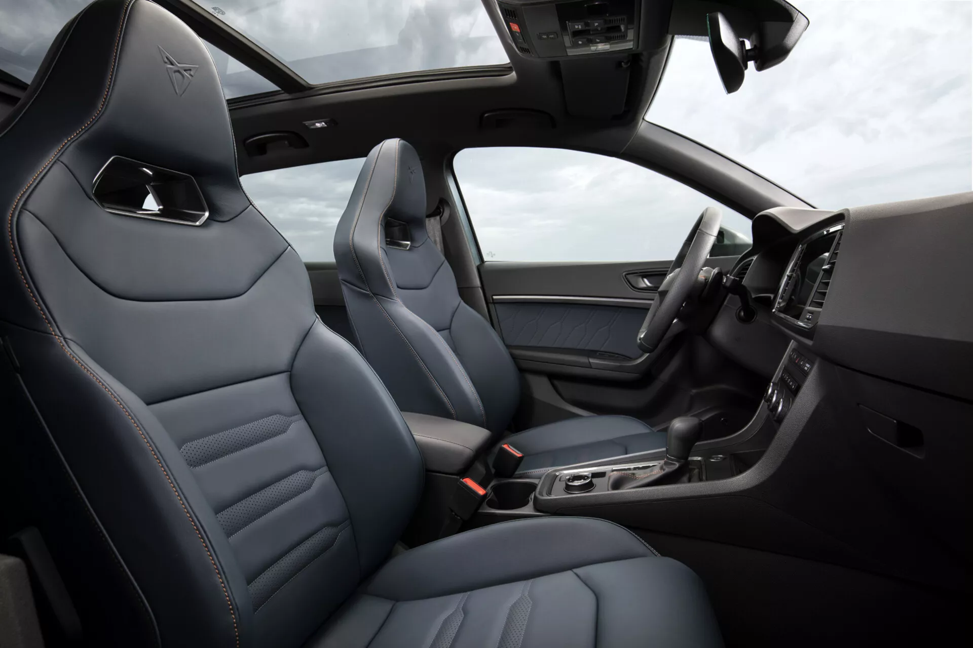 Vue intérieure du Cupra Ateca 2020, montrant des sièges en cuir de luxe avec coutures en diamant, console centrale élégante avec fonctionnalités technologiques, affichage numérique du tableau de bord et partie du volant avec commandes, sous un toit ouvrant ensoleillé