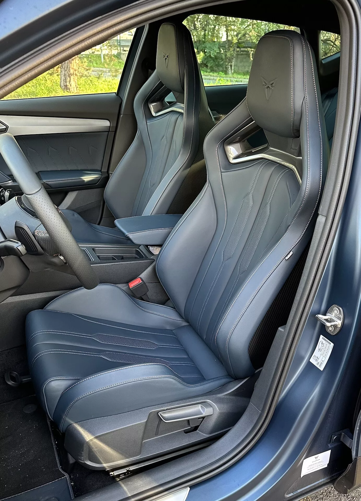 Vue intérieure d'un Cupra Formentor 2020 montrant le siège du conducteur depuis une perspective de trois quarts, avec la porte ouverte, sièges baquets sportifs avec coutures bleu clair, et un aperçu de la console centrale et du tableau de bord.