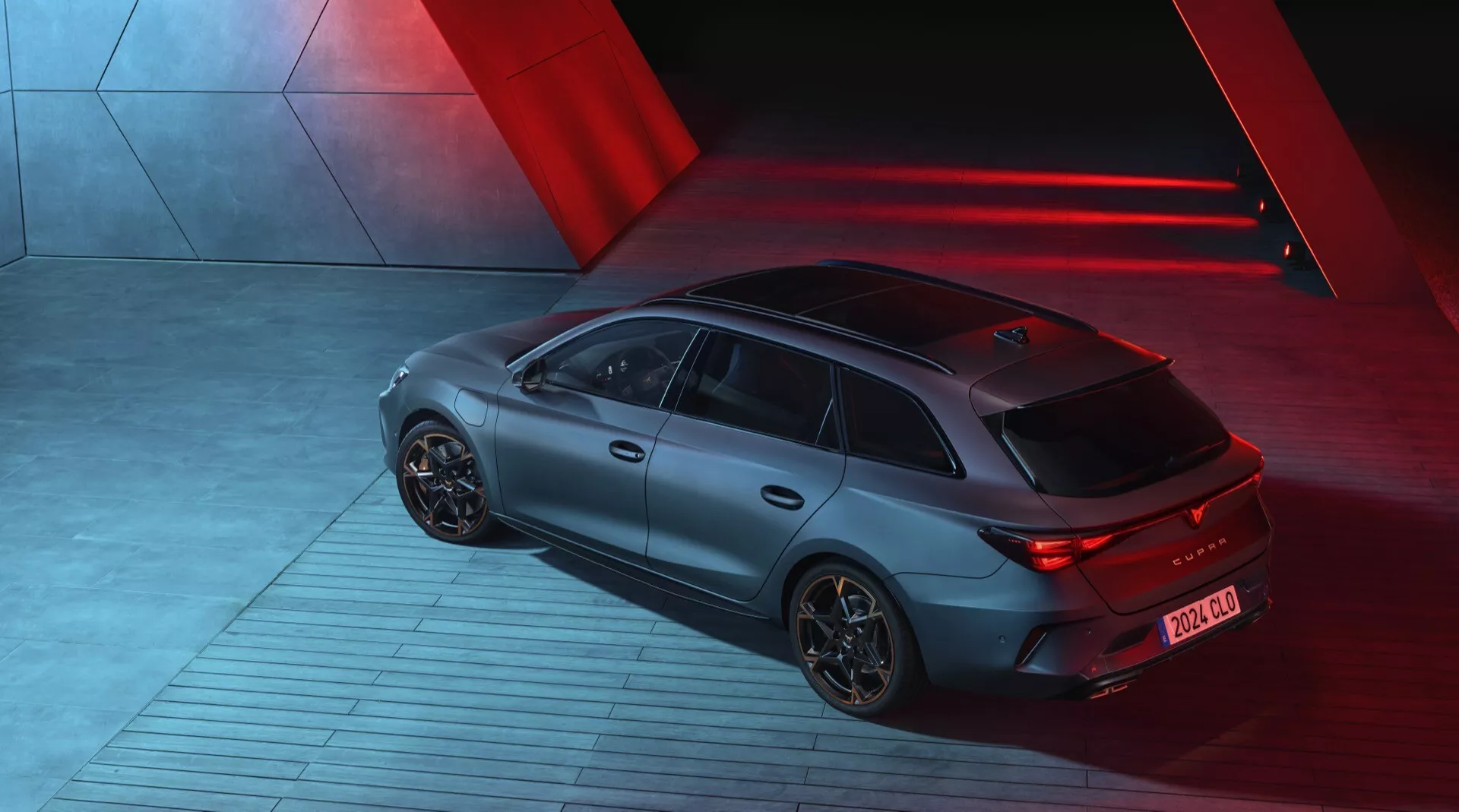 Vue en hauteur de la Cupra Leon 2024 (1ère génération restylée) prise de nuit, montrant l'arrière et le côté droit du sportback. La voiture est éclairée par des lumières directionnelles rouges dramatiques créant une ambiance élégante et futuriste. Elle présente une finition mate, des feux arrière à LED illuminés et des jantes en alliage aérodynamiques. Le véhicule est stationné sur une surface industrielle à motifs, suggérant un mouvement. Les caractéristiques technologiques visibles incluent un diffuseur arrière et une antenne courte à l'arrière pour les technologies de communication.