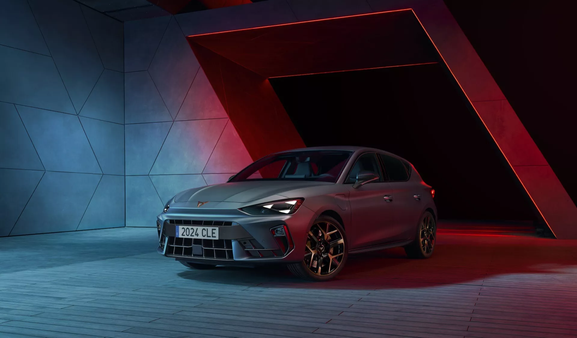 Image de la Cupra Leon 2024 (1ère génération restylée) dans un cadre de studio dynamique avec un arrière-plan rouge et bleu frappant. La voiture est capturée sous un angle de trois quarts avant bas, mettant en valeur la calandre avant, les phares à LED, les jantes alliage, le design aérodynamique, la finition de peinture avancée et les poignées de porte distinctives. La composition et l'éclairage mettent en avant les lignes élégantes de la carrosserie et les caractéristiques de design du véhicule.