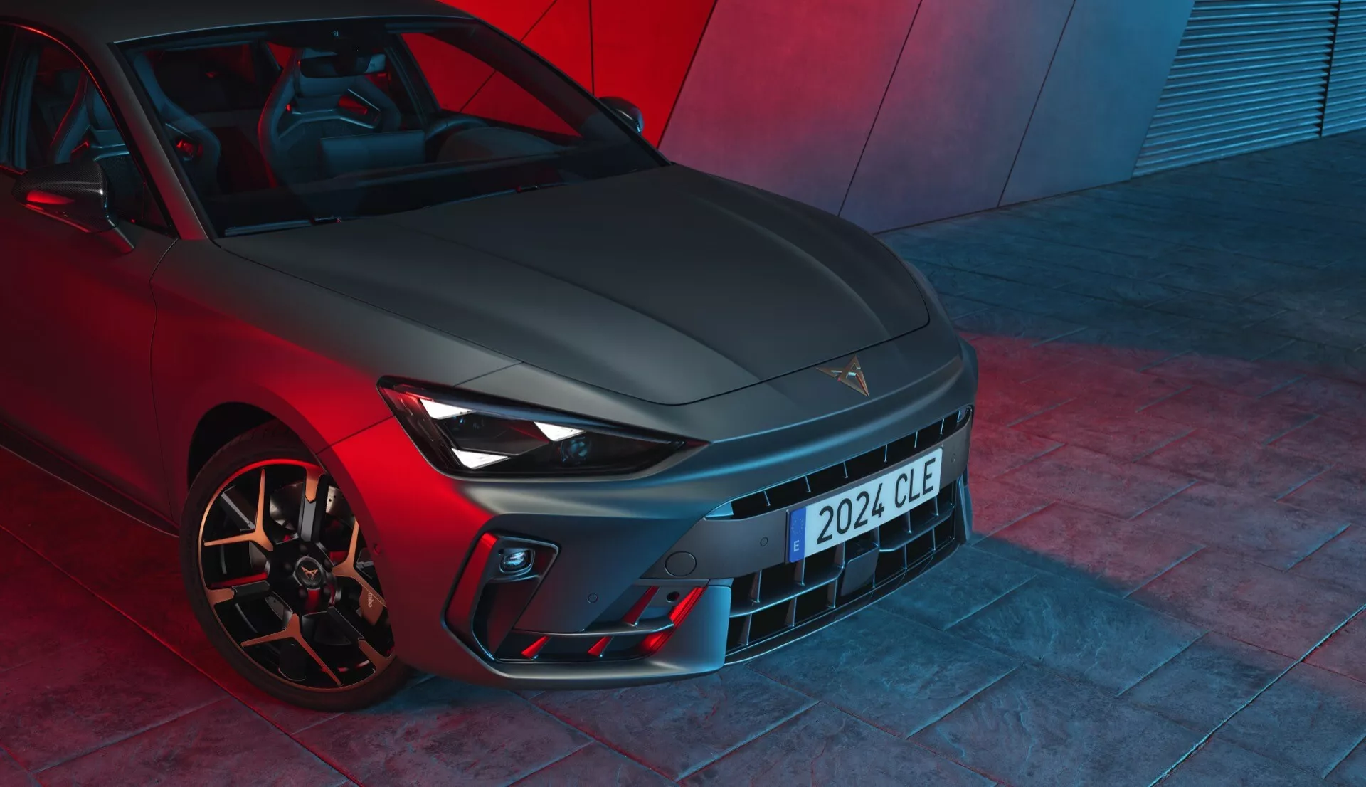 Vue de face basse de la Cupra Leon 2024 (première génération avec lifting), mettant en valeur ses caractéristiques de design avec une calandre proéminente, des phares élégants dotés de feux de jour à LED, un pare-chocs sportif avec des prises d'air et des roues en alliage axées sur les performances avec des étriers de frein haute performance. La finition métallique de la peinture et l'éclairage ambiant rouge dramatique suggèrent un véhicule haut de gamme axé sur les performances, probablement photographié sur le sol d'un showroom.