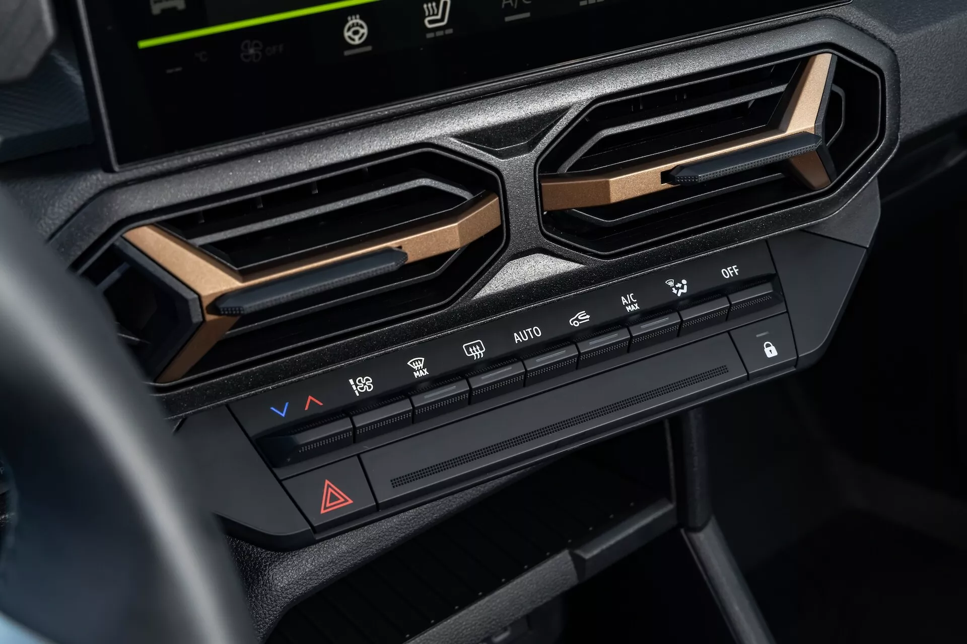 Vue rapprochée du système de contrôle climatique sur la Dacia Duster de 3ème génération 2024 sur le tableau de bord central sous l'écran d'infotainment, mettant en lumière les réglages de température et de vitesse du ventilateur, les fonctions de distribution de l'air, les caractéristiques de dégivrage, le bouton des feux de détresse, et les options de climatisation, encadrées par des grilles d'aération angulaires et accentuées avec une finition métallique pour une esthétique intérieure moderne
