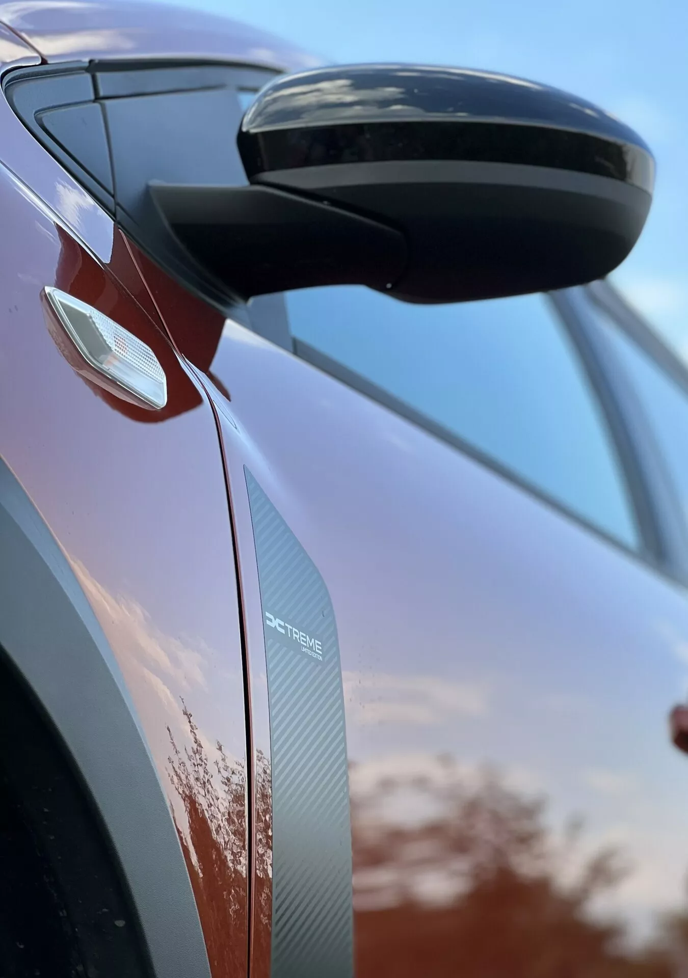Détail en gros plan du Dacia Jogger 2022 se concentrant sur le rétroviseur latéral et le design de la porte, présentant des caractéristiques aérodynamiques, des clignotants intégrés et des textures en fibre de carbone sur fond de ciel