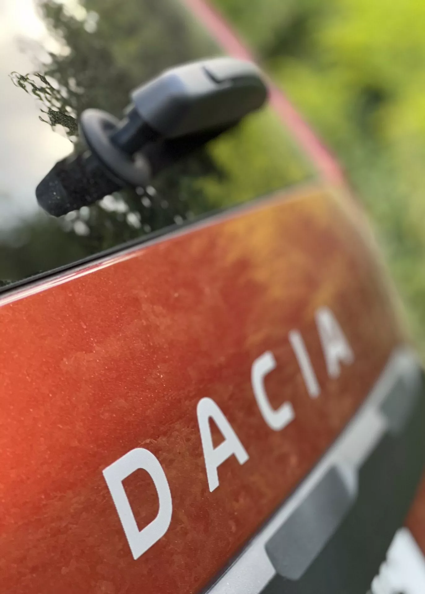 Vue rapprochée du panneau arrière du Dacia Jogger 2022 avec le nom DACIA en lettres capitales blanches sur une peinture métallisée orange-rouge avec des taches et de la poussière, suggérant une utilisation et une durabilité en extérieur, située dans un environnement naturel avec un rétroviseur flou indiquant des caractéristiques modernes du miroir.