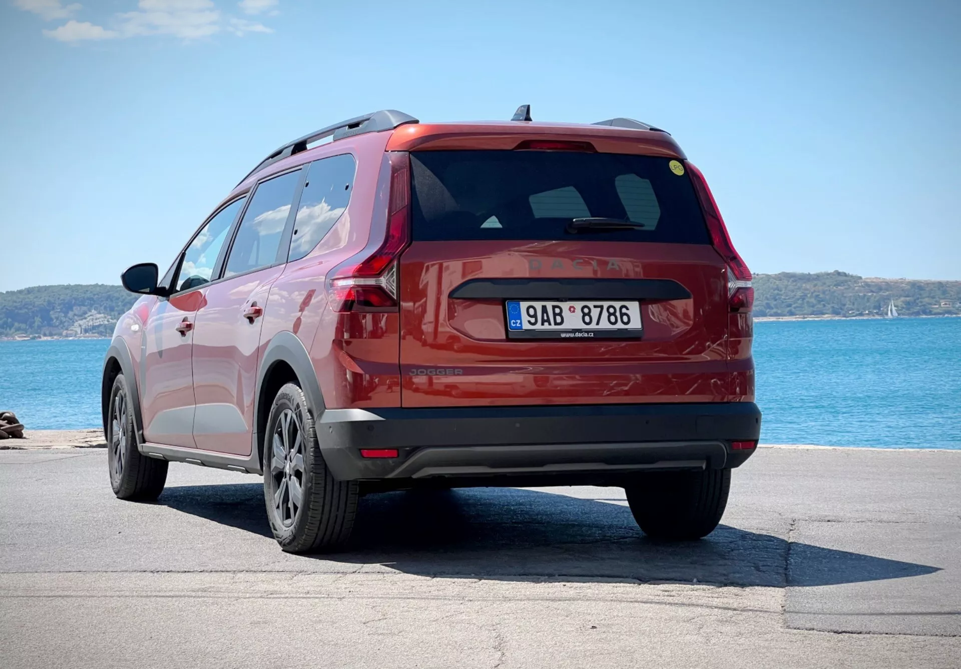 Dacia Jogger 2022 en rouge, capturée depuis un angle arrière de trois quarts montrant le côté et la conception du hayon du SUV, garée près d'un plan d'eau avec l'horizon en arrière-plan, mettant en lumière ses jantes en alliage à multiples branches, ses feux arrière LED et son aileron arrière aérodynamique