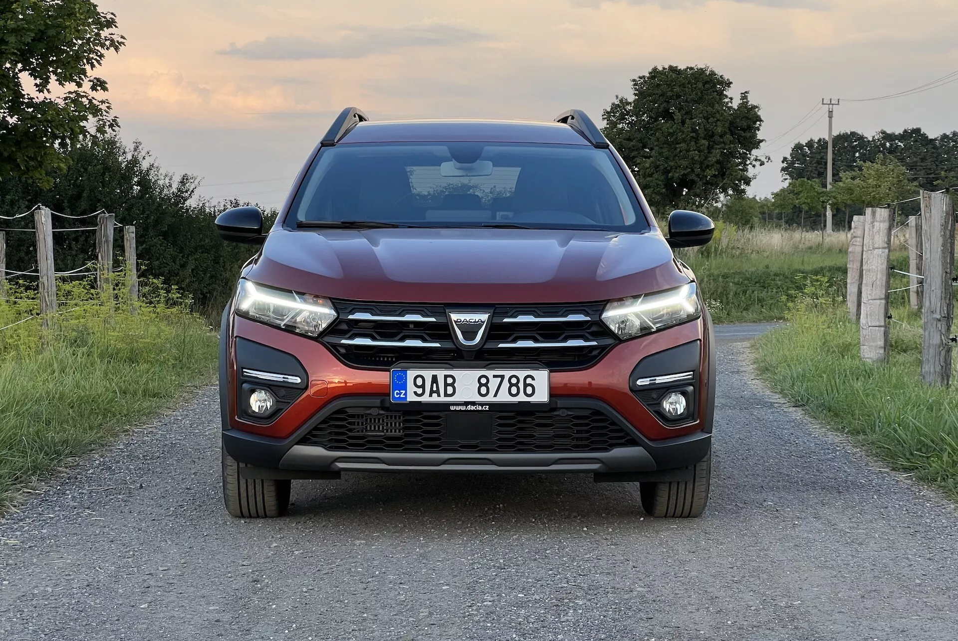Dacia Jogger 2022 positionnée sur une route rurale étroite, offrant une vue frontale symétrique qui met en évidence le design moderne de la voiture, y compris une grille avant proéminente avec un grand emblème, des phares LED élégants, des lignes de capot profilées, un pare-chocs avant avec feux de brouillard et capteurs de stationnement, et des rétroviseurs latéraux avec clignotants intégrés.
