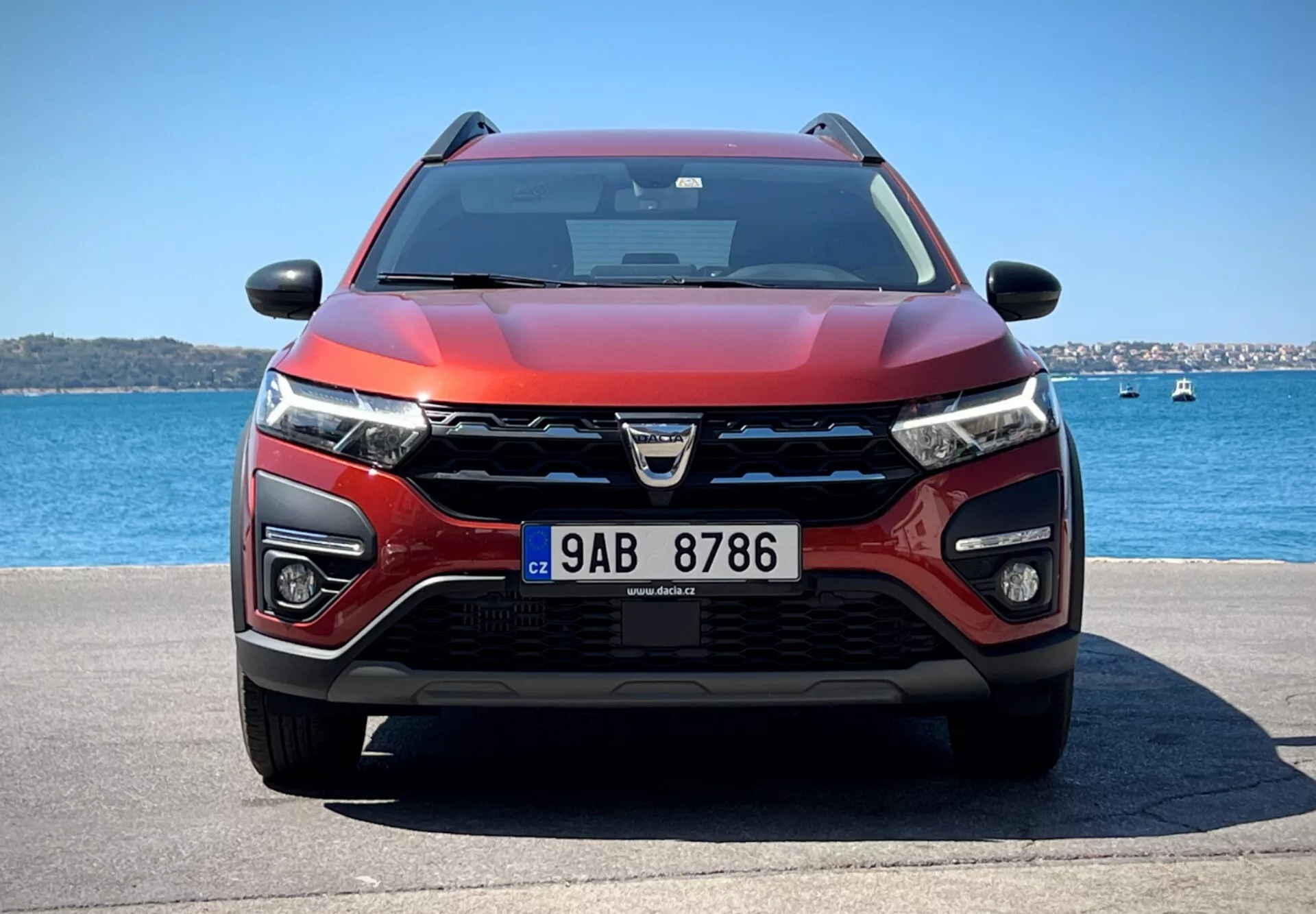 Dacia Jogger 2022 exposant son design moderne de crossover et ses caractéristiques technologiques avancées dans un cadre aquatique serein, avec une grille proéminente portant le logo, des phares LED, des feux de brouillard, des capteurs ADAS potentiels, des prises d'air pour le refroidissement et le radar, un schéma de couleurs rouge et noir bicolore, un capot aérodynamique, équipé d'essuie-glaces indiquant une possible technologie sensible à la pluie, des rétroviseurs avec clignotants intégrés pouvant contenir une technologie de surveillance des angles morts, sous un ciel clair.