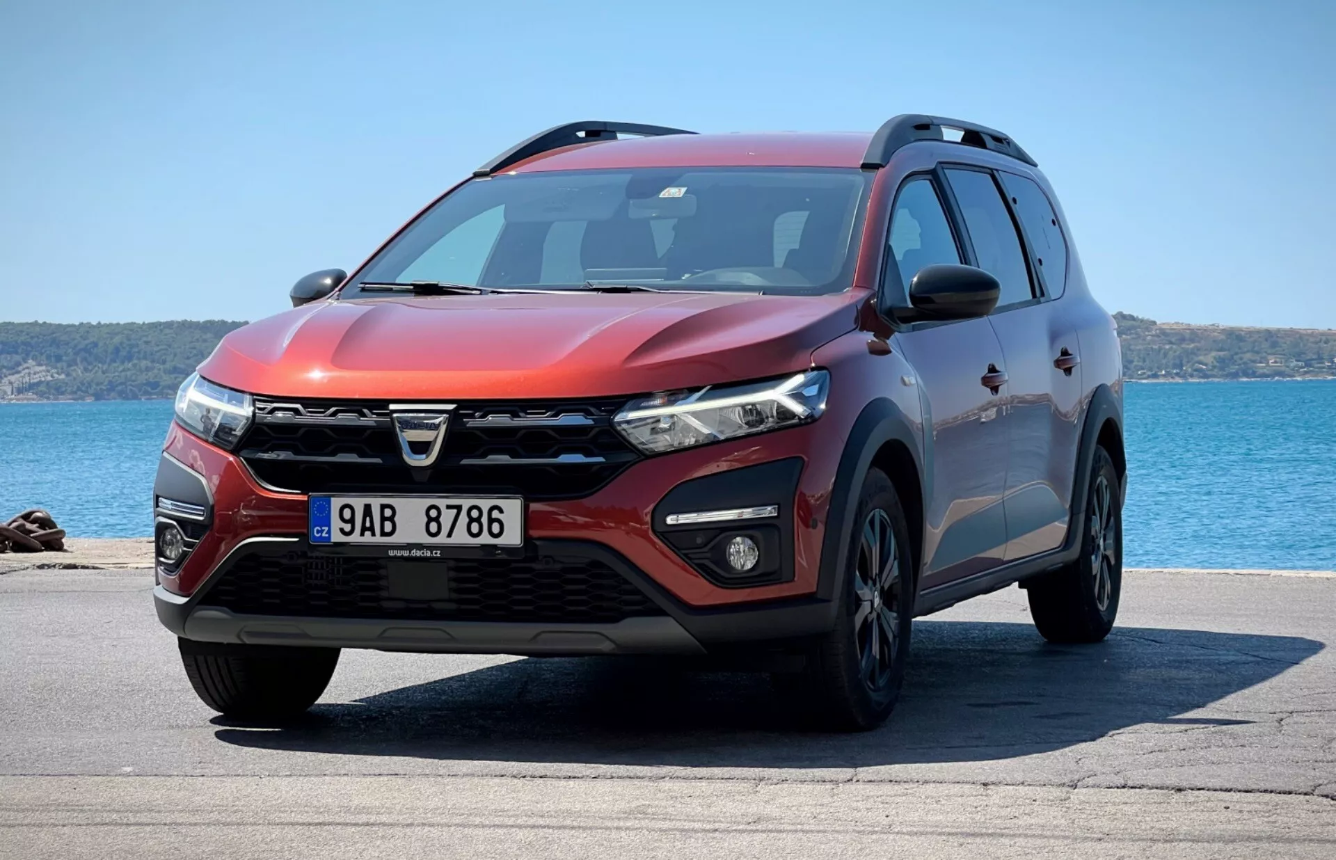 Vue de trois quarts avant du SUV Dacia Jogger 2022 garé avec un arrière-plan océanique, montrant un design de grille distinct, des phares LED, des feux de brouillard, un pare-chocs avant aérodynamique avec prises d'air et capteurs de stationnement, des contours de capot musculaires, des roues en alliage, des clignotants intégrés sur les rétroviseurs latéraux, un profil latéral avec protection inférieur, des barres de toit, des vitres teintées et une extérieur rouge métallique sous un soleil éclatant indiquant le profil de design, les caractéristiques technologiques et l'utilité.