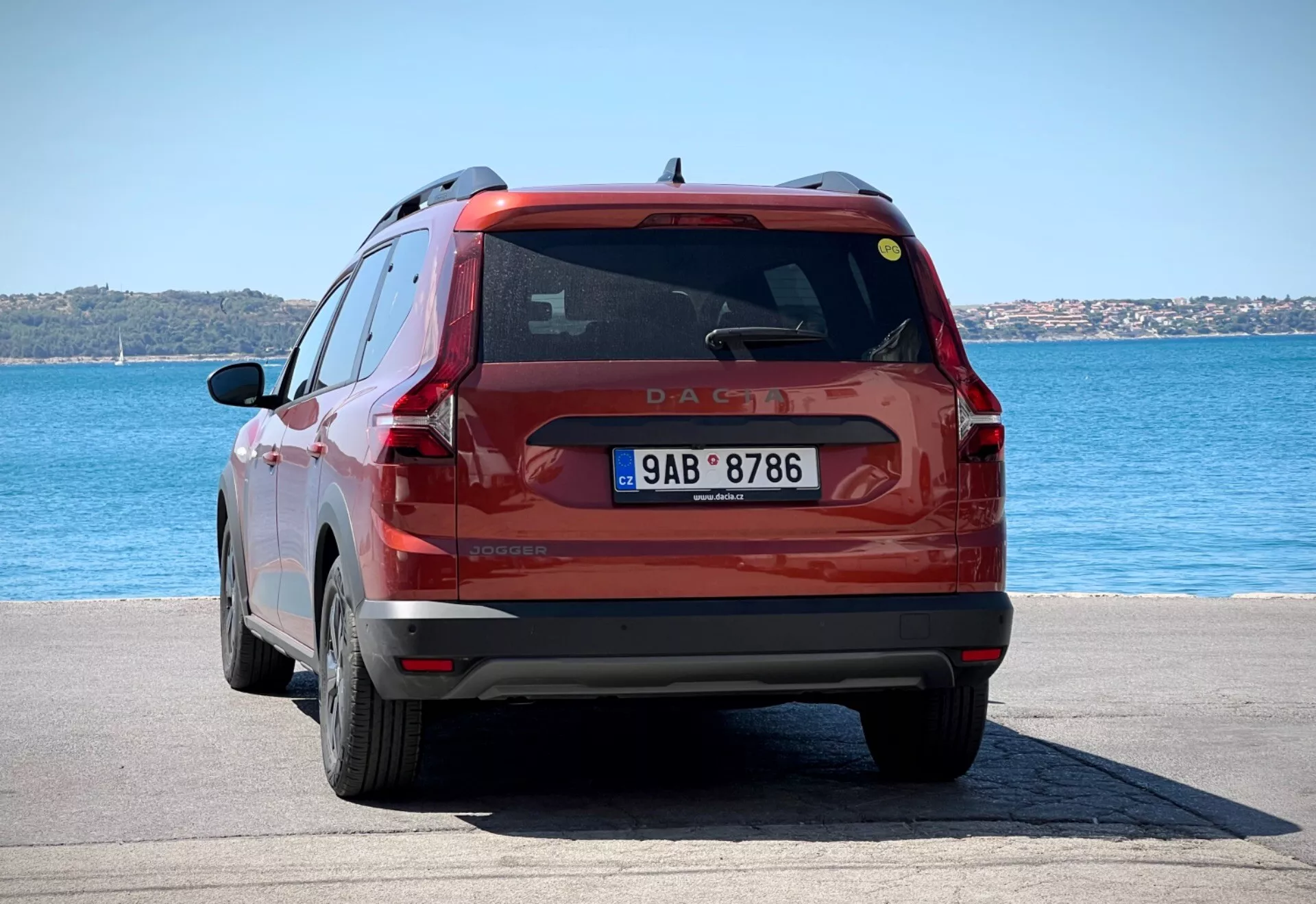 Vue arrière d'un SUV compact rouge Dacia Jogger 2022 garé sur une zone pavée au bord de l'eau sous la lumière vive du jour, montrant l'ensemble de l'arrière du véhicule, y compris les feux arrière, la lunette arrière et la plaque d'immatriculation sur un fond d'eau bleue et de cieux clairs.