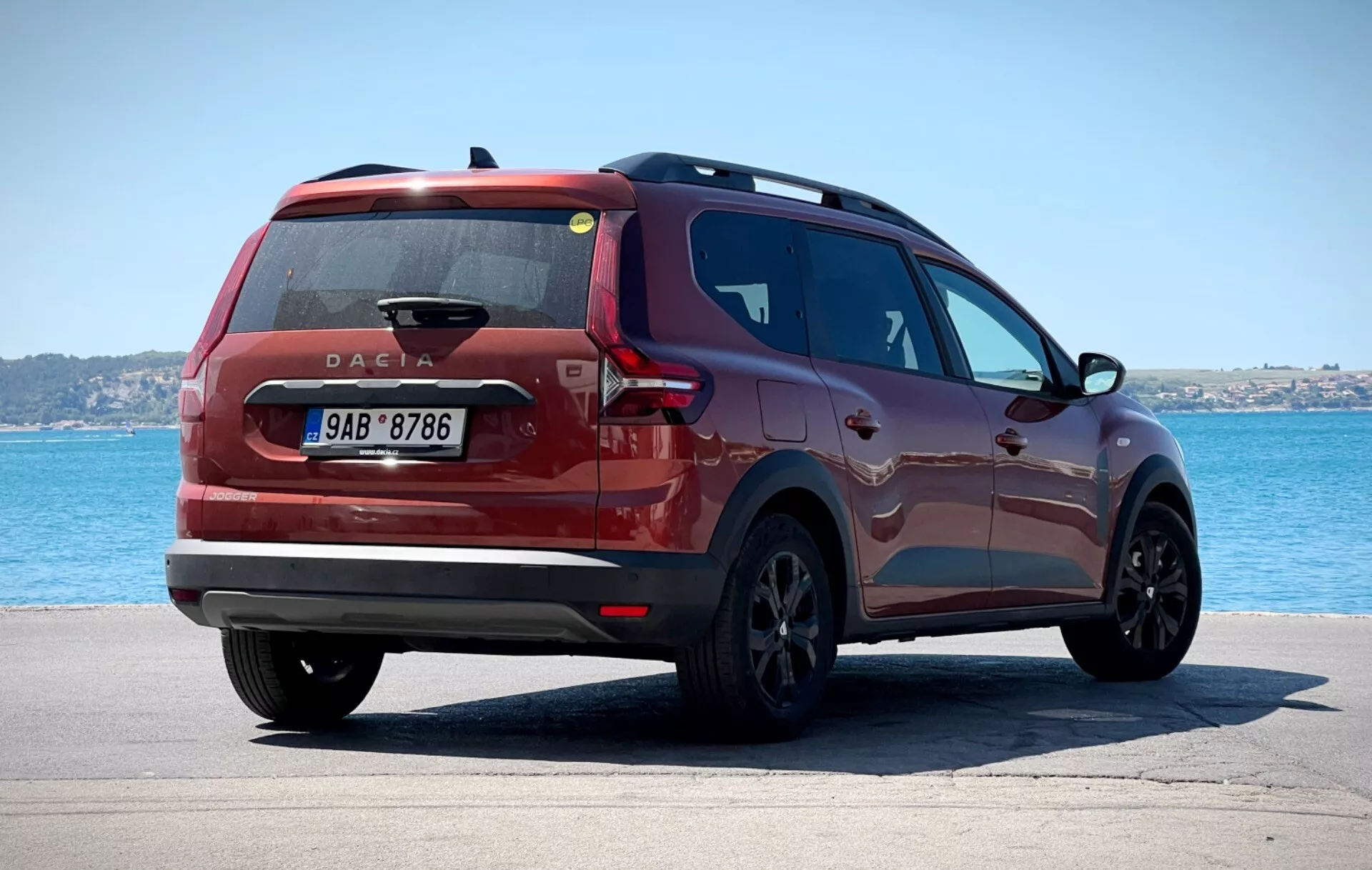 Dacia Jogger 2022 photographiée sous un angle arrière de trois quarts positionnée sur une surface pavée avec un plan d'eau et un ciel dégagé en arrière-plan, affichant des feux arrière modernes, un insigne de Dacia placé au centre et un profil latéral élégant contre un environnement côtier ou lacustre tranquille.