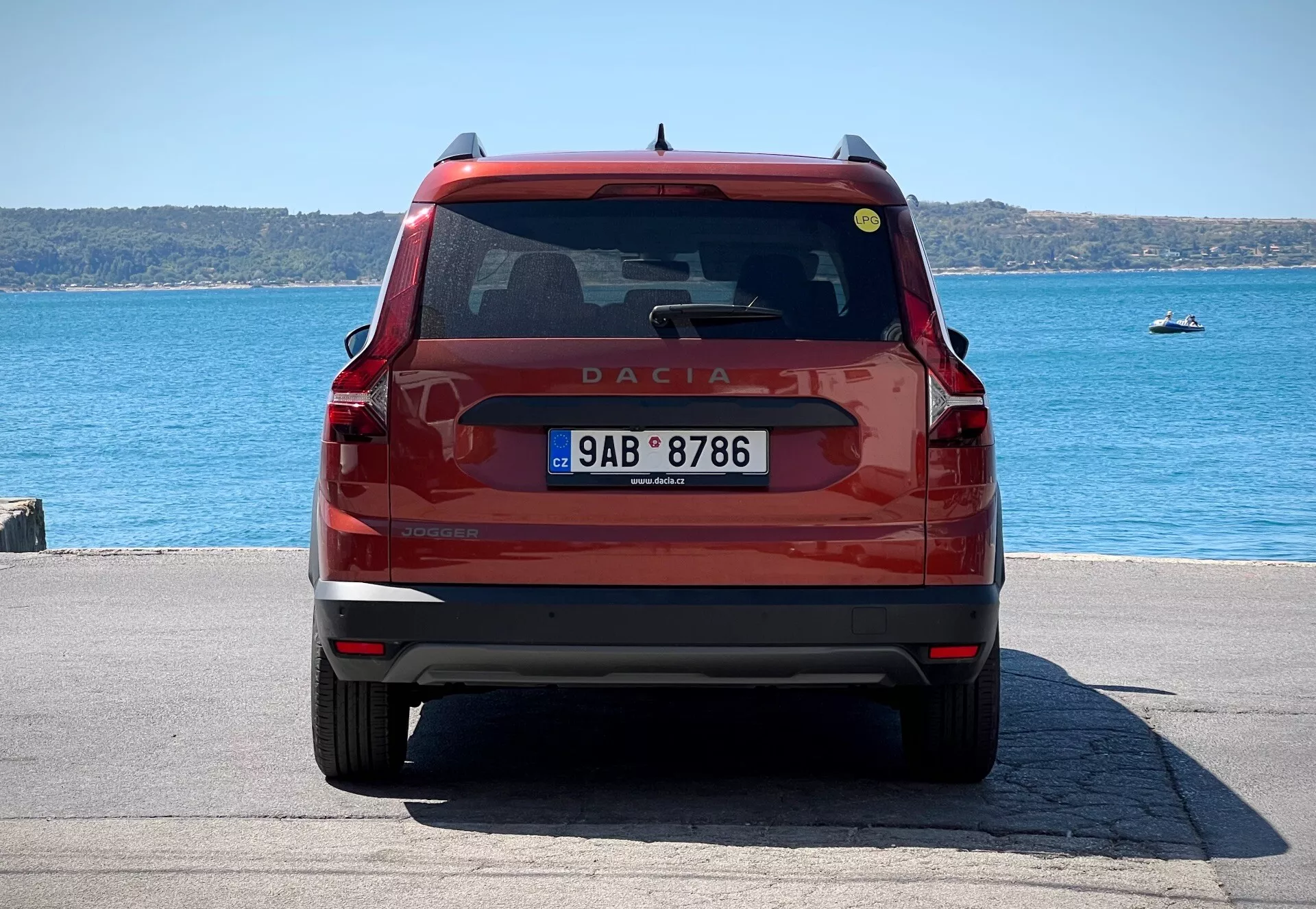 Vue arrière d'une Dacia Jogger 2022 rouge, montrant le hayon avec l'emblème et le nom du modèle, le pare-brise arrière avec essuie-glace et feu de stop, des feux arrière modernes, la plaque d'immatriculation et le pare-chocs avec réflecteurs et potentiels capteurs de stationnement, garée au bord de l'eau en plein jour.