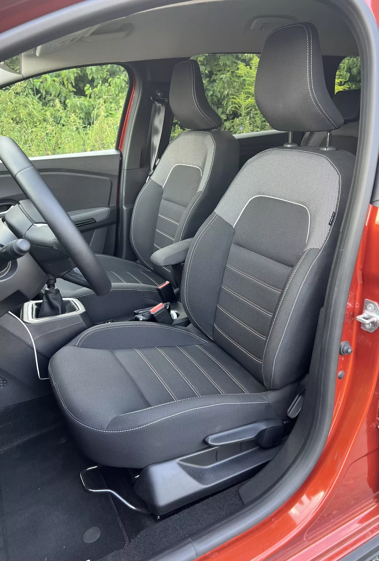 Vue détaillée de l'intérieur du Dacia Jogger 2022 montrant la zone passager avant avec des sièges noirs garnis de motifs linéaires, des commandes d'ajustement, une partie de la console centrale et un aperçu de l'extérieur rouge