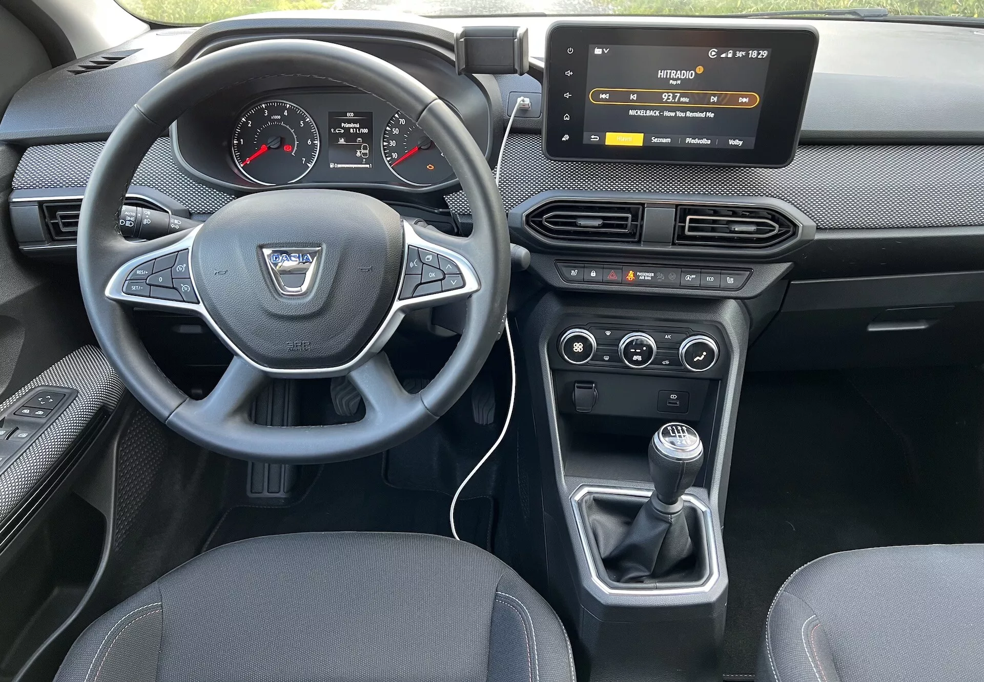 Vue intérieure d'une Dacia Jogger 2022 depuis le siège arrière, soulignant le tableau de bord du conducteur, le volant avec logo du fabricant et boutons de commande, le combiné d'instrumentation digital-analogique, le système d'infodivertissement tactile, les commandes HVAC, le contrôle climatique bi-zone et le levier de vitesse manuel