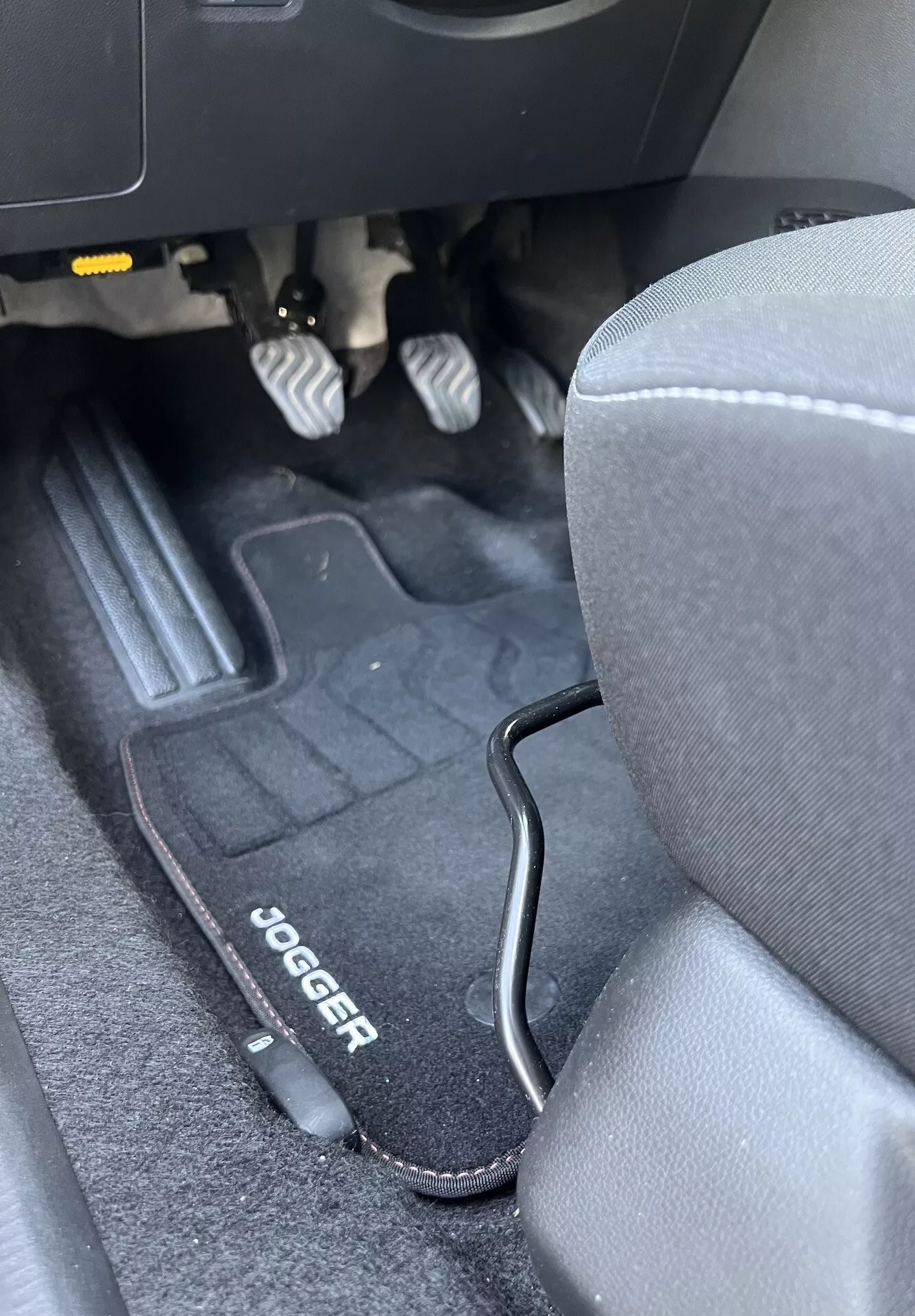 Intérieur de la Dacia Jogger 2022 montrant l'espace pour les pieds côté conducteur avec pédales métalliques, tapis de sol marqué, siège en tissu gris, et garniture en plastique noir