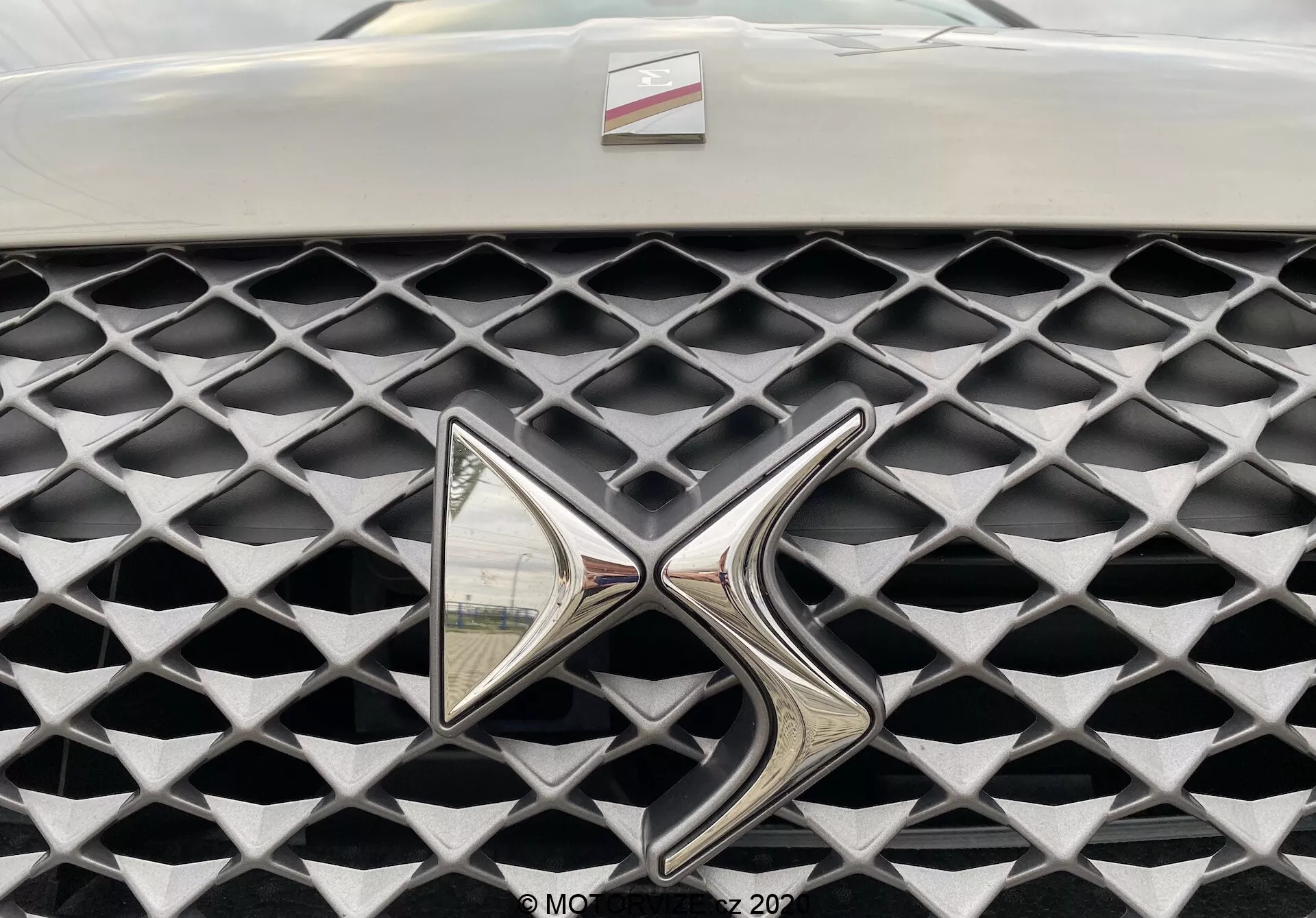 Gros plan sur la calandre de la DS Automobiles DS 3 Crossback E-Tense 2019. L'image capture la calandre sous un angle frontal, mettant l'accent sur un emblème métallique très réfléchissant. La calandre présente une texture caractéristique de cellules en forme de diamant avec une finition grise mate, contrastant avec l'emblème chromé brillant. Champ de vision limité au-delà de la calandre et de l'emblème, avec un arrière-plan flou.