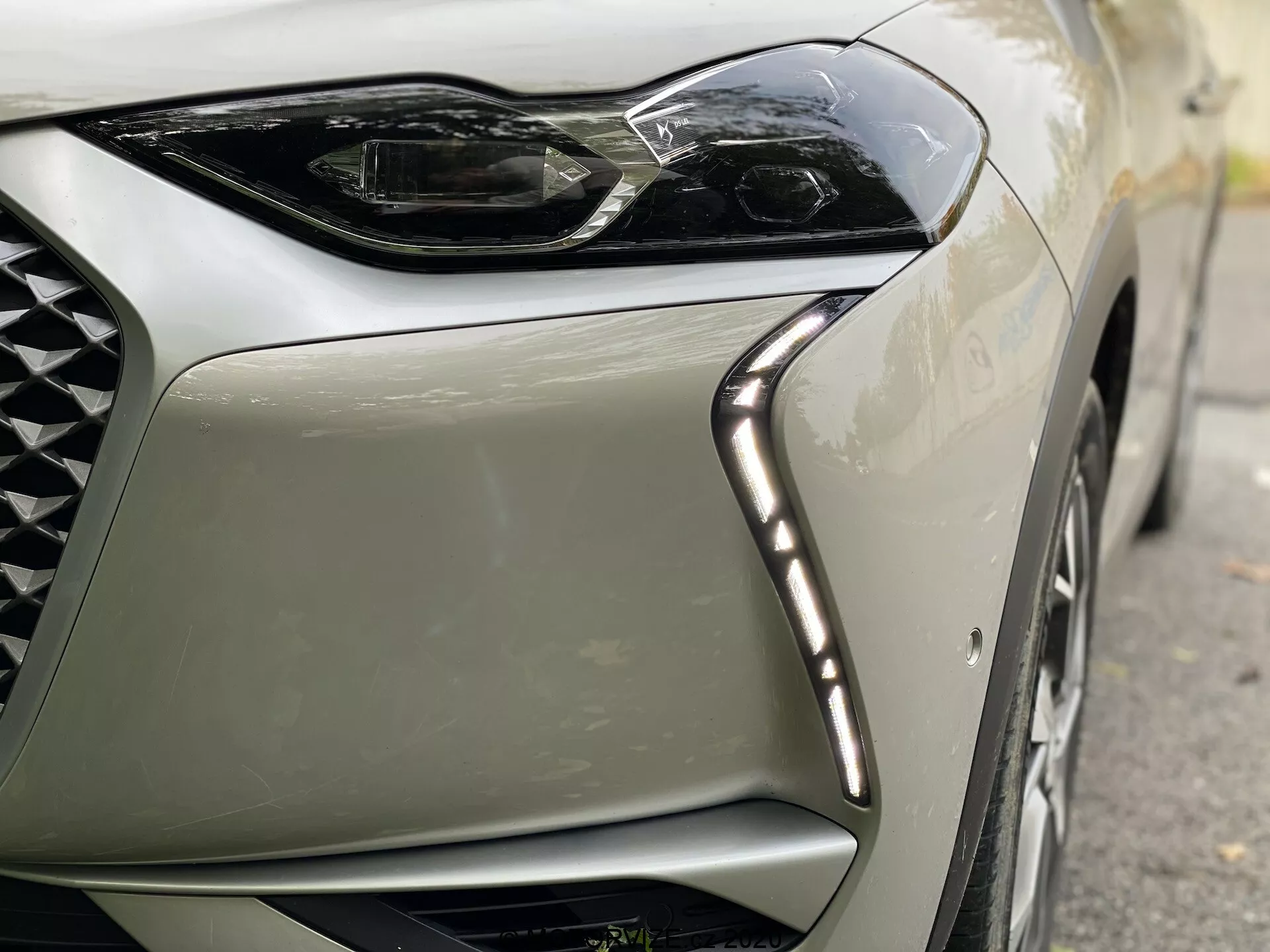 Vue rapprochée du coin avant gauche d'une voiture moderne DS Automobiles DS-3 Crossback E-Tense de 2019. L'image se concentre sur le design angulaire et élégant des phares à LED, avec des feux de jour à LED le long du bord supérieur et une partie de la grille en maille de diamant, soulignant le luxe et la technologie de pointe.