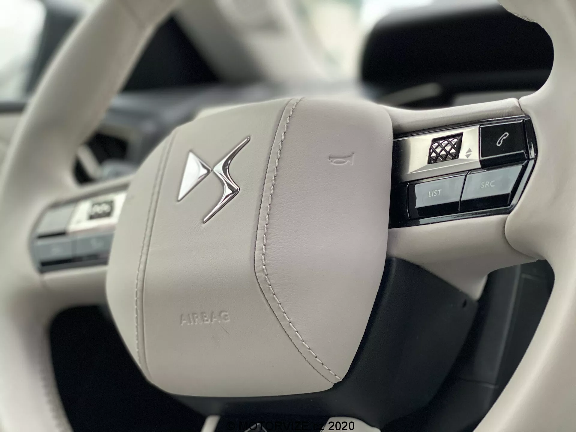 Gros plan sur le côté gauche d'un volant moderne recouvert de cuir clair du DS Automobiles DS 3 Crossback E-Tense 2019, mettant en avant l'artisanat et le luxe avec des coutures visibles. Comprend plusieurs boutons intégrés pour le contrôle des fonctions du véhicule, y compris la commande vocale, le téléphone, le menu d'accueil et les commandes multimédia, ainsi qu'un emblème central proéminent et un boîtier à l'inscription 'AIRBAG'.
