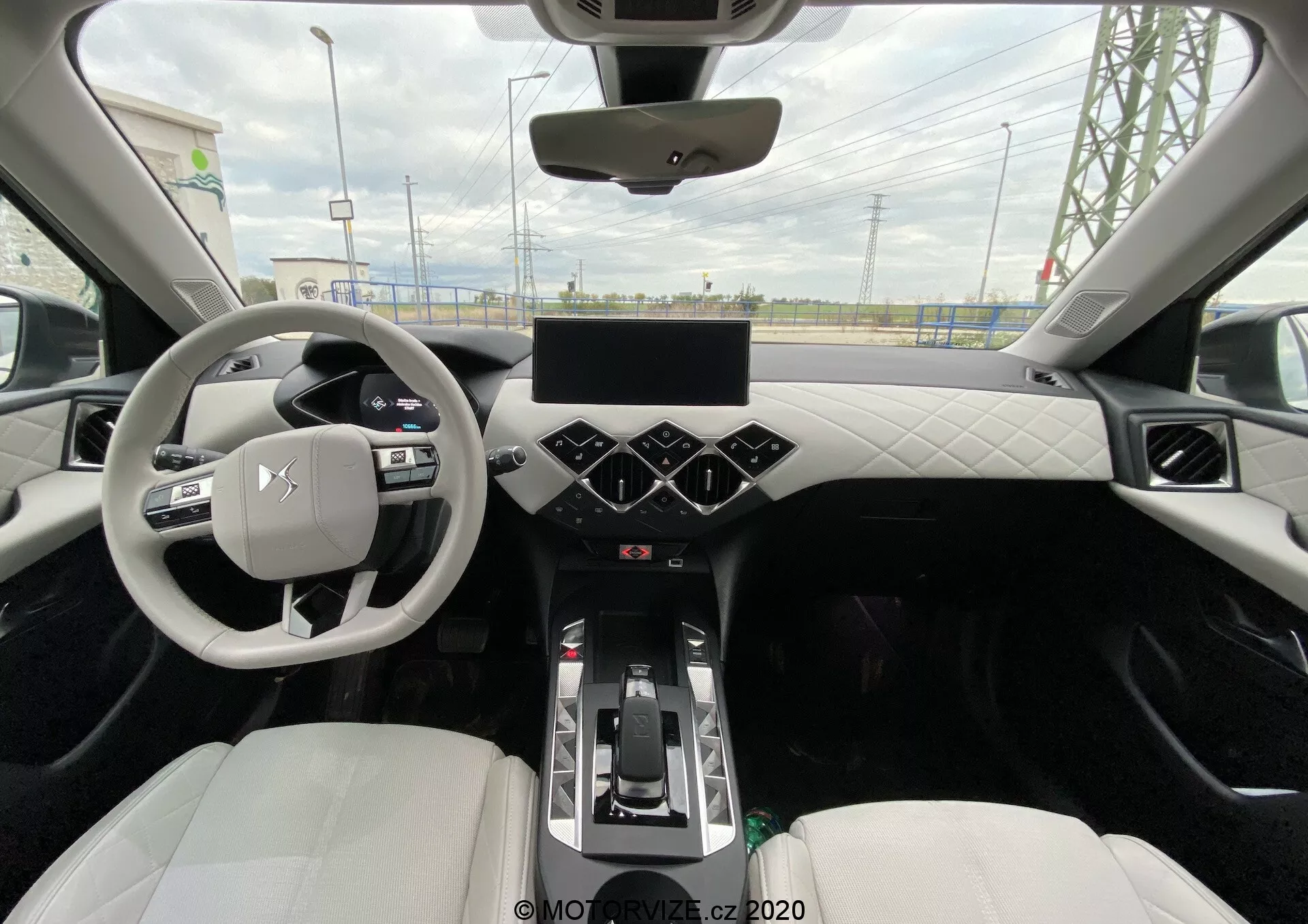 Vue depuis le siège arrière d'une DS Automobiles DS-3 Crossback E-Tense 2019 montrant l'intérieur central moderne. La photo capture un volant à trois branches avec commandes intégrées, un tableau de bord numérique, un grand écran tactile sur la console centrale avec des boutons hexagonaux de contrôle climatique en dessous et un levier de vitesses indiquant une transmission automatique. Le tableau de bord et les portes présentent un design capitonné utilisant du cuir ou de la similicuir, et les sièges baquets avant sont habillés de cuir ou de matériau synthétique de couleur claire. Les caractéristiques notables incluent un éclairage d'ambiance le long du pourtour, un toit panoramique et des aérations visibles sur le tableau de bord.