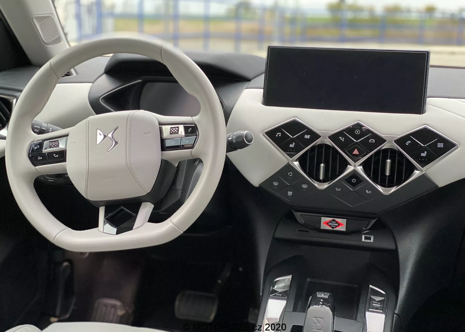 Vue intérieure d'une DS Automobiles DS 3 Crossback E-Tense 2019 avec un volant argenté/gris clair centré avec boutons de contrôle et logo du fabricant, à travers lequel on aperçoit le siège du conducteur. Le tableau de bord noir présente des éléments géométriques avec des aérateurs centraux triangulaires, un écran d'infodivertissement éteint et en dessous, de possibles commandes de climatisation et tunnel central. Sur la gauche, une vue partielle de la porte du conducteur avec des commandes de fenêtres et rétroviseurs.
