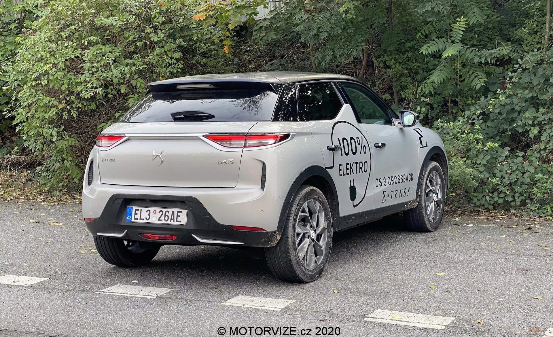 Automobile DS Automobiles DS 3 2019 (DS 3 Crossback E-Tense) capturée sous un angle trois quarts arrière, mettant en évidence le profil arrière et latéral du véhicule. Les caractéristiques de conception incluent des feux arrière orientés horizontalement, une garniture chromée, des poignées de porte affleurantes, un port de charge et des roues en alliage à plusieurs branches, symbolisant une voiture électrique moderne. La voiture est garée en plein air dans un environnement urbain ou suburbain sous un ciel couvert, avec des arbres à feuilles caduques à proximité.
