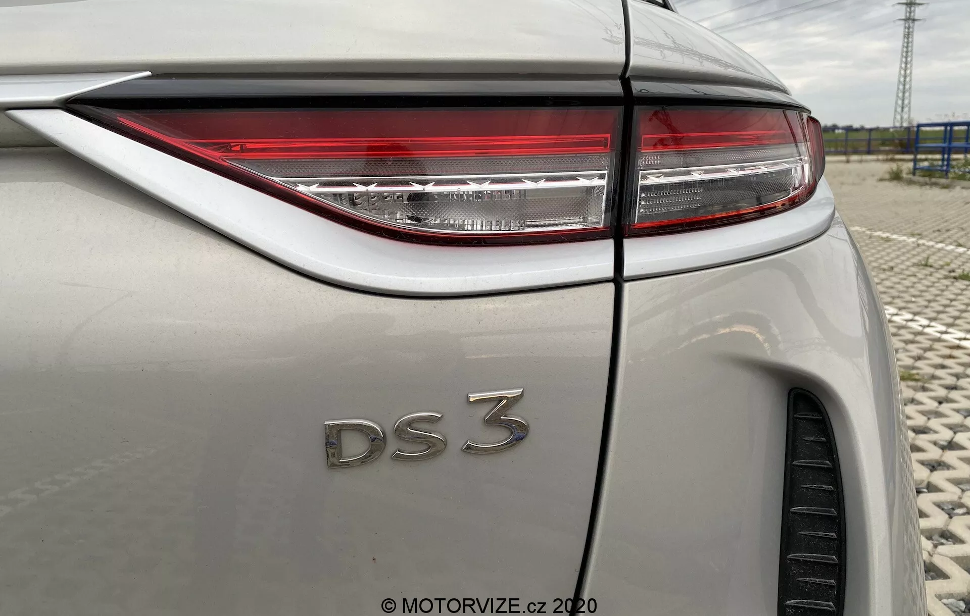 Vue rapprochée en trois-quarts arrière de la DS Automobiles DS 3 Crossback E-Tense 2019. L'image se concentre sur le côté droit du véhicule, mettant en évidence les feux arrière LED nets, le badge 'DS 3' et une carrosserie métallique légère. Les caractéristiques visibles incluent la ligne de pli du garde-boue arrière, une partie du pneu arrière, l'antenne en forme d'aileron de requin et la transition de la vitre arrière dans la ligne de toit, le tout sous un reflet de ciel nuageux.