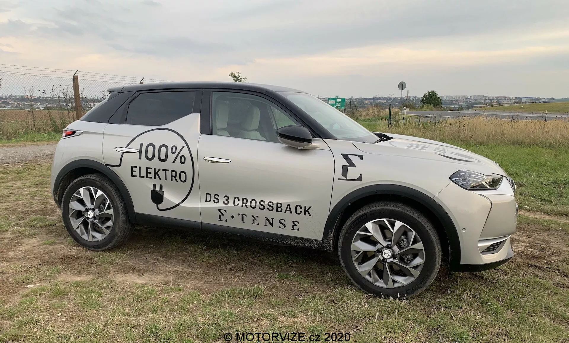 Vue de profil latéral d'un SUV moderne, le DS 3 Crossback E-Tense de 2019, un véhicule électrique avec des graphismes '100% ELEKTRO' sur le côté. Positionné sur un sol herbeux avec un bout de route pavée en arrière-plan, permettant une vue claire de la calandre avant, des phares LED, de la roue avant et du profil sous un léger angle. Les caractéristiques visibles comprennent des jantes en alliage multi-rayons, des rétroviseurs latéraux avec indicateurs intégrés, des poignées de porte affleurantes et un symbole de port de charge électrique sur l'aile avant, suggérant un accent sur la technologie avancée, le style et l'écologie, le tout sous un éclairage naturel et nuageux.