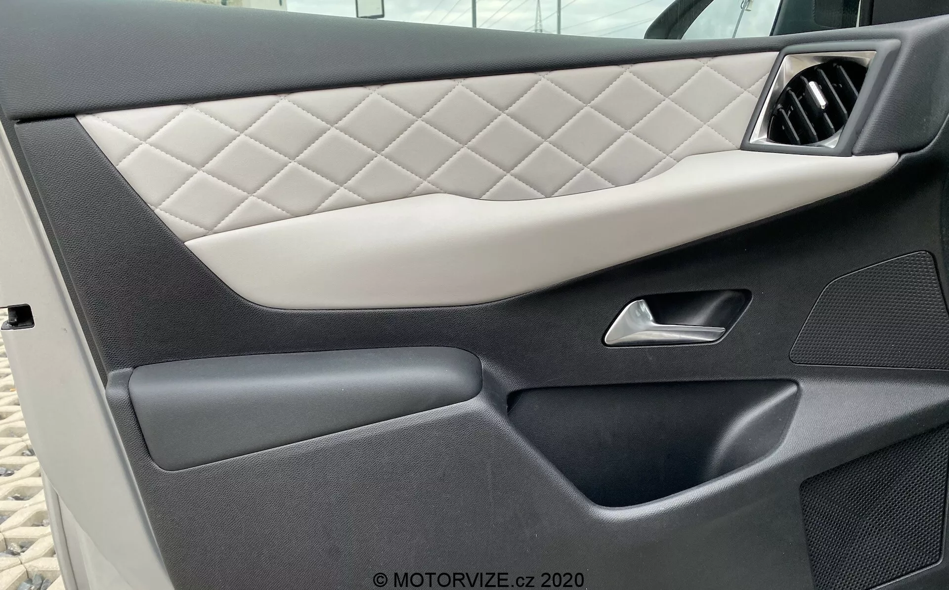 Vue rapprochée du panneau de porte intérieur du véhicule DS Automobiles DS-3 2019 (DS-3-Crossback-E-Tense), présentant une sellerie matelassée à motif losange, une poignée argentée élégante, des sections en plastique foncé de haute qualité avec des textures variées et une poche de rangement inférieure intégrée, mettant en valeur le design luxueux et détaillé de la voiture.