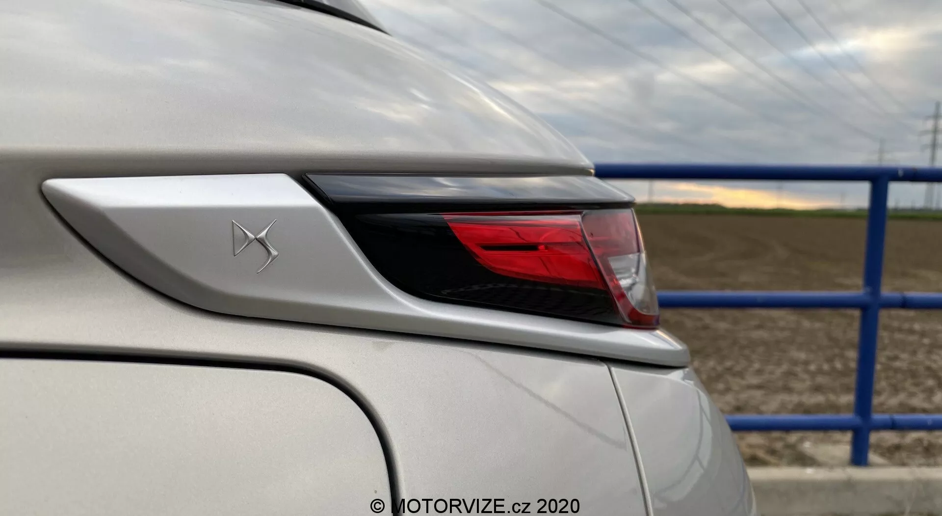 Gros plan du coin arrière d'un véhicule blanc DS-3 2019 (DS-3 Crossback E-Tense), centré sur les éléments de design et les caractéristiques autour de la zone du feu arrière. La photo prise de l'angle arrière gauche met en avant la courbure du feu arrière et de l'aile arrière, montrant les détails de la carrosserie et l'assemblage avancé des feux à LED. Un emblème avec un X stylisé est visible au-dessus du feu arrière, avec la peinture blanche en parfait état reflétant une finition brillante. À l'arrière-plan, il y a une route avec une clôture métallique bleue et un champ agricole.