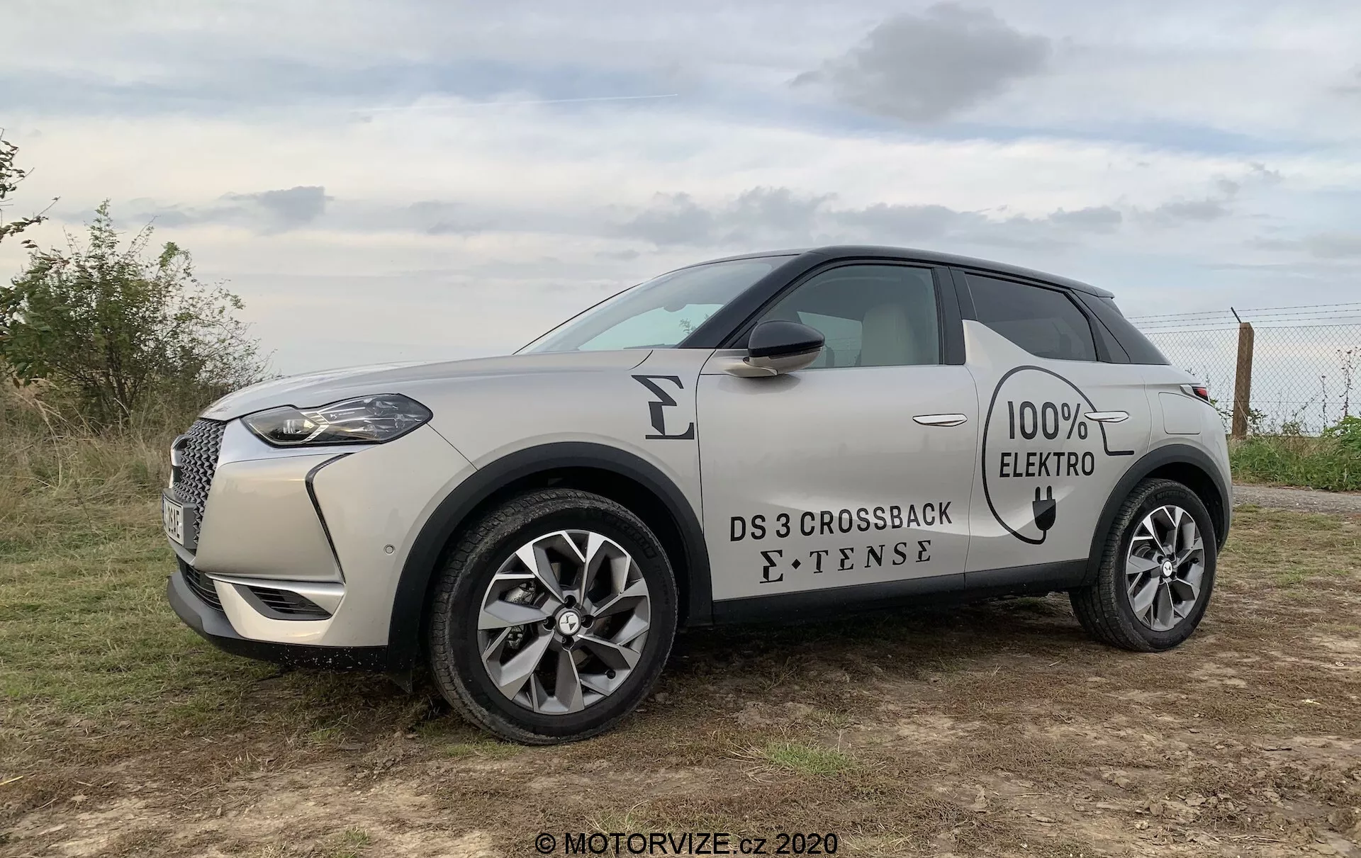 Profil latéral d'un SUV compact électrique DS 3 Crossback E-Tense 2019 dans une teinte sable claire avec un toit noir contrastant. L'image capture la calandre avant avec cadre chromé, les phares à LED, les badges '100% ELECTRO' et 'E-TENSE' sur le côté, les jantes en alliage à plusieurs branches et les poignées de porte affleurantes. Le véhicule est positionné à l'extérieur sur un terrain herbeux sous un ciel couvert, suggérant un éclairage de début de matinée ou de fin d'après-midi.