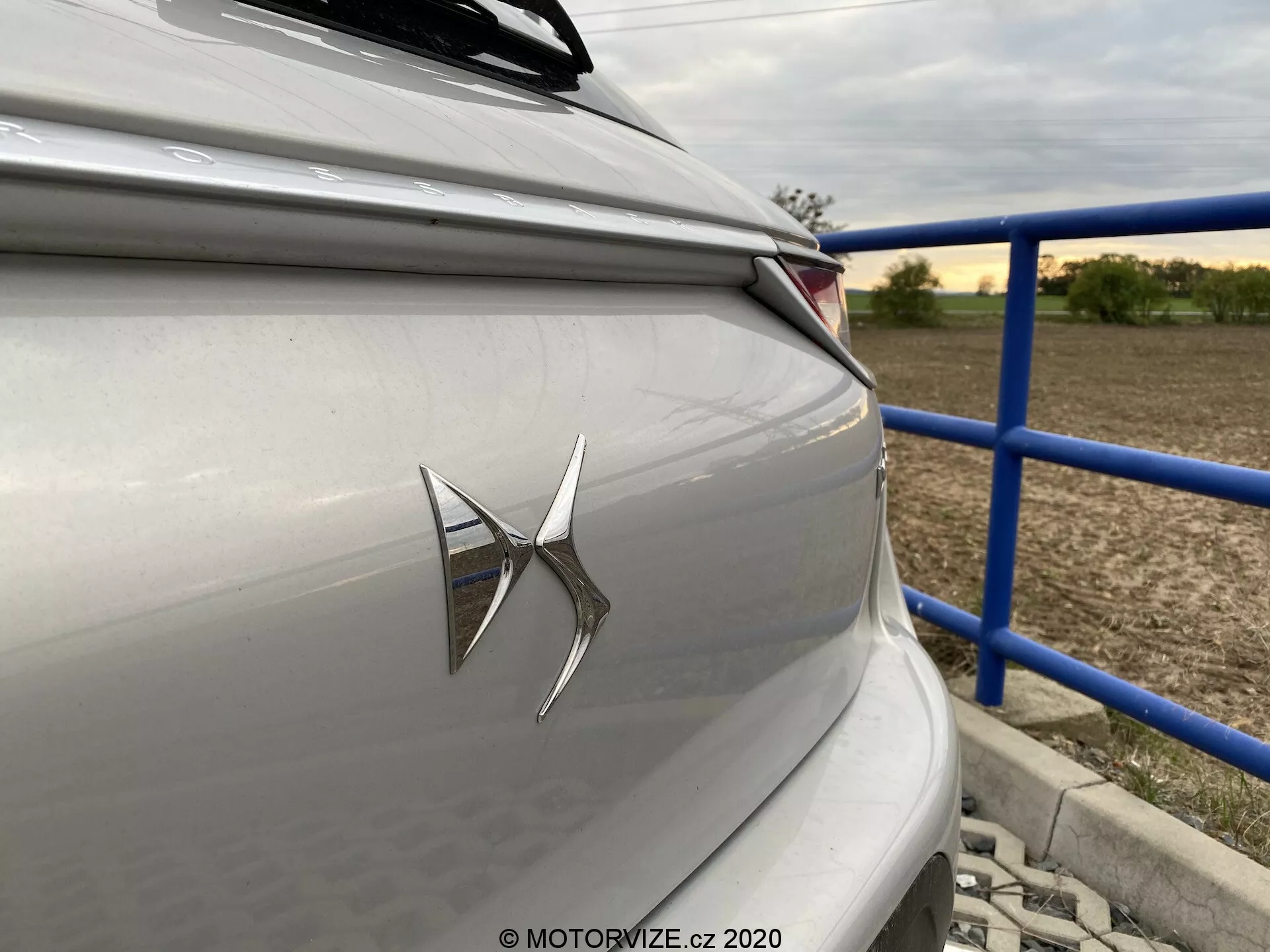Gros plan sur la section arrière d'une berline ou d'une hatchback de luxe de couleur argent clair ou gris, avec un emblème de marque proéminent sur le coffre et un groupe de feux arrière LED modernes. La photographie, prise sous un léger angle depuis l'arrière droit du véhicule, montre des lignes de design horizontales nettes et un arrière-plan suburbain ou rural avec des rampes bleues et des structures. L'esthétique du design de la voiture met en avant la technologie contemporaine et un schéma de couleur neutre et sobre.