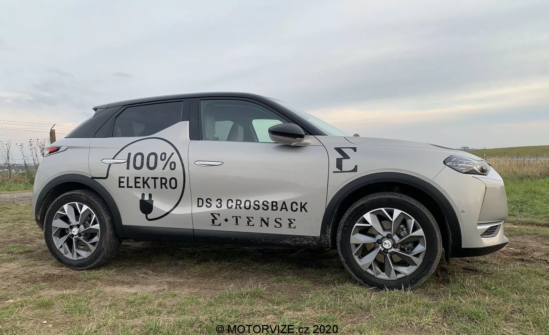 Profil latéral du DS Automobiles DS 3 Crossback E-Tense 2019, un véhicule électrique, pris sous un léger angle avant, mettant en avant la roue avant, la porte côté conducteur et le rétroviseur, avec des vues partielles de la calandre avant et de la roue arrière, positionné sur un champ herbeux avec un arrière-plan subtil.
