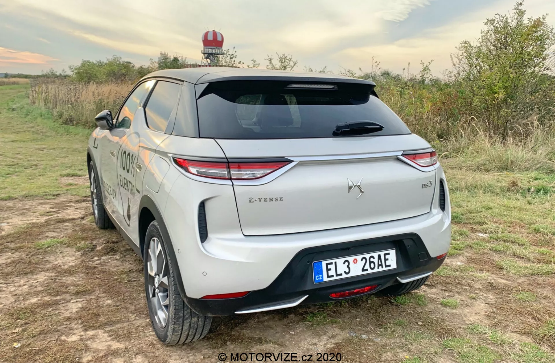 Vue trois quarts arrière d'un SUV DS Automobiles DS-3 Crossback E-Tense 2019 garé sur une surface non pavée dans un cadre rural, présentant des feux arrière à LED, un emblème bien en vue, un essuie-glace arrière, un pare-chocs arrière distinctif avec capteurs, une finition bicolore, une plaque d'immatriculation centrale, un badge de niveau de finition, un porte-bagages, une éventuelle lumière d'urgence, une ligne de carrosserie aérodynamique lisse, des roues en alliage, des rétroviseurs latéraux de la couleur de la carrosserie et un clapet de port de charge indiquant qu'il s'agit d'un véhicule électrique ou hybride rechargeable.