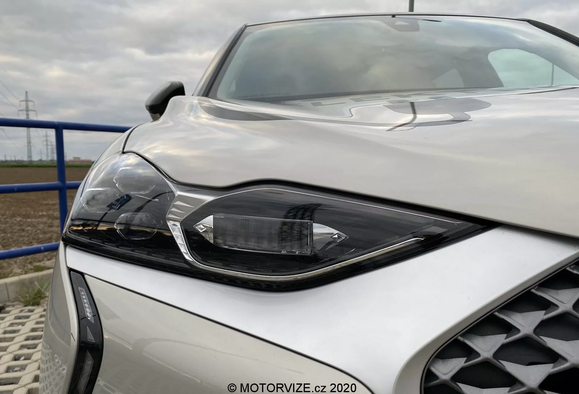 Gros plan de la partie avant de la DS Automobiles DS-3 Crossback E-Tense 2019 sous un angle bas et légèrement décentré vers la droite, mettant en valeur le phare avant droit avec une technologie LED ou HID angulaire et élégante présentant des segments distincts pour le faisceau principal, le faisceau élevé et éventuellement les feux de jour. Les lignes sculptées du capot et la calandre en nid d'abeille sont également visibles, mettant en avant le design sportif et luxueux du véhicule argent métallisé.