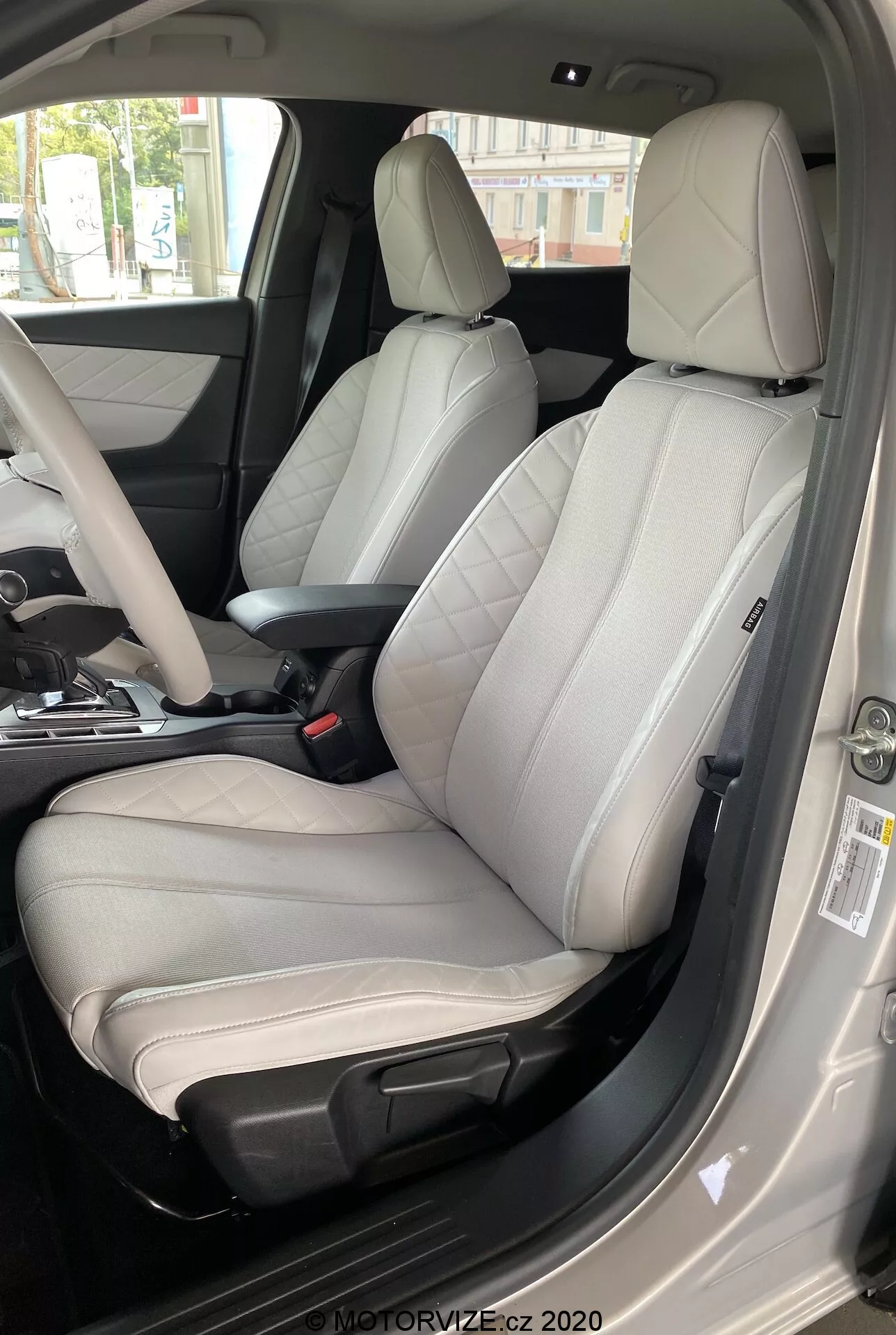Intérieur de la DS Automobiles DS 3 Crossback E-Tense 2019, se concentrant sur le siège du conducteur depuis une perspective extérieure côté conducteur. L'image, prise d'un angle bas vers haut, présente un siège en cuir bicolore moderne avec un centre gris clair et des bords gris foncé ou noirs. Clairement conçu pour le confort et l'esthétique sportive, le siège dispose de renforts latéraux proéminents, de coutures visibles et de commandes de réglage électronique. La porte du conducteur ouverte, la ceinture de sécurité visible, la console centrale partiellement visible et le panneau de porte avec divers boutons de commande indiquent des matériaux haut de gamme et de nombreuses fonctionnalités avancées. L'éclairage diurne améliore la clarté des détails intérieurs luxueux.