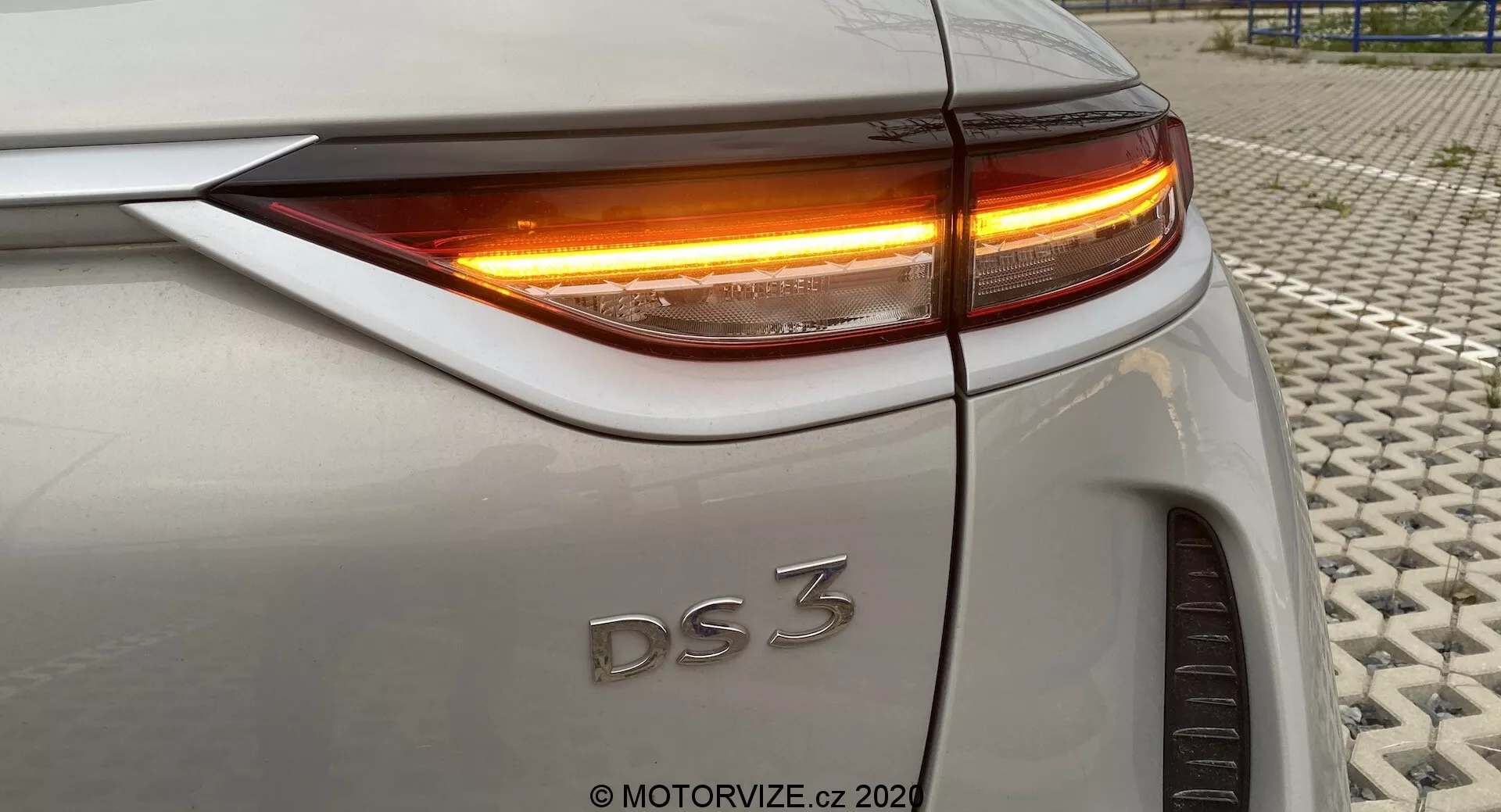 Gros plan de la partie arrière d'une DS 3 Crossback E-Tense (modèle 2019), montrant le quart arrière droit sous un angle légèrement élevé. L'image met en avant le feu arrière LED et le clignotant avec une illumination orange vif. Les éléments visibles comprennent le bord supérieur du coffre, la courbure de l'aile arrière, l'alignement précis des panneaux de carrosserie et une partie de la roue arrière droite. La voiture présente une peinture argentée ou gris clair. Photo prise sur une surface texturée.