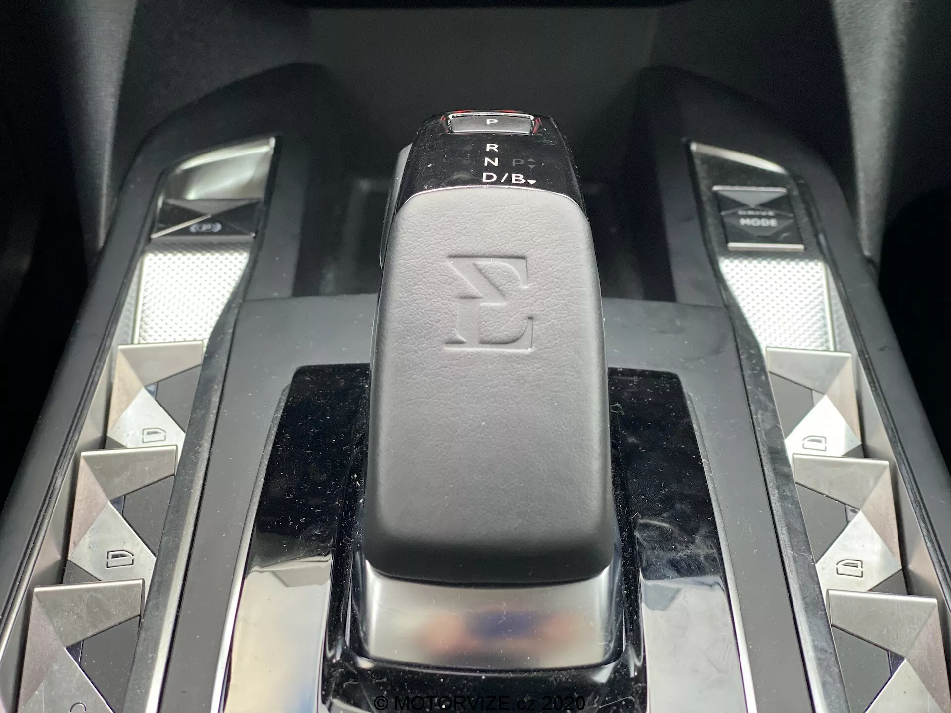 Gros plan sur le levier de vitesses d'une DS Automobiles DS-3 2019 (DS-3 Crossback E-Tense), capturé sous un angle légèrement surélevé de face. Le levier de vitesses, avec une surface en cuir ou en similicuir noir avec des indicateurs PRND, est au centre de l'image. Des boutons symétriques en forme de losange argenté et deux curseurs métalliques encadrent le levier, suggérant des fonctionnalités supplémentaires de la voiture. Les matériaux hautement brillants et haut de gamme dégagent un intérieur moderne et luxueux.
