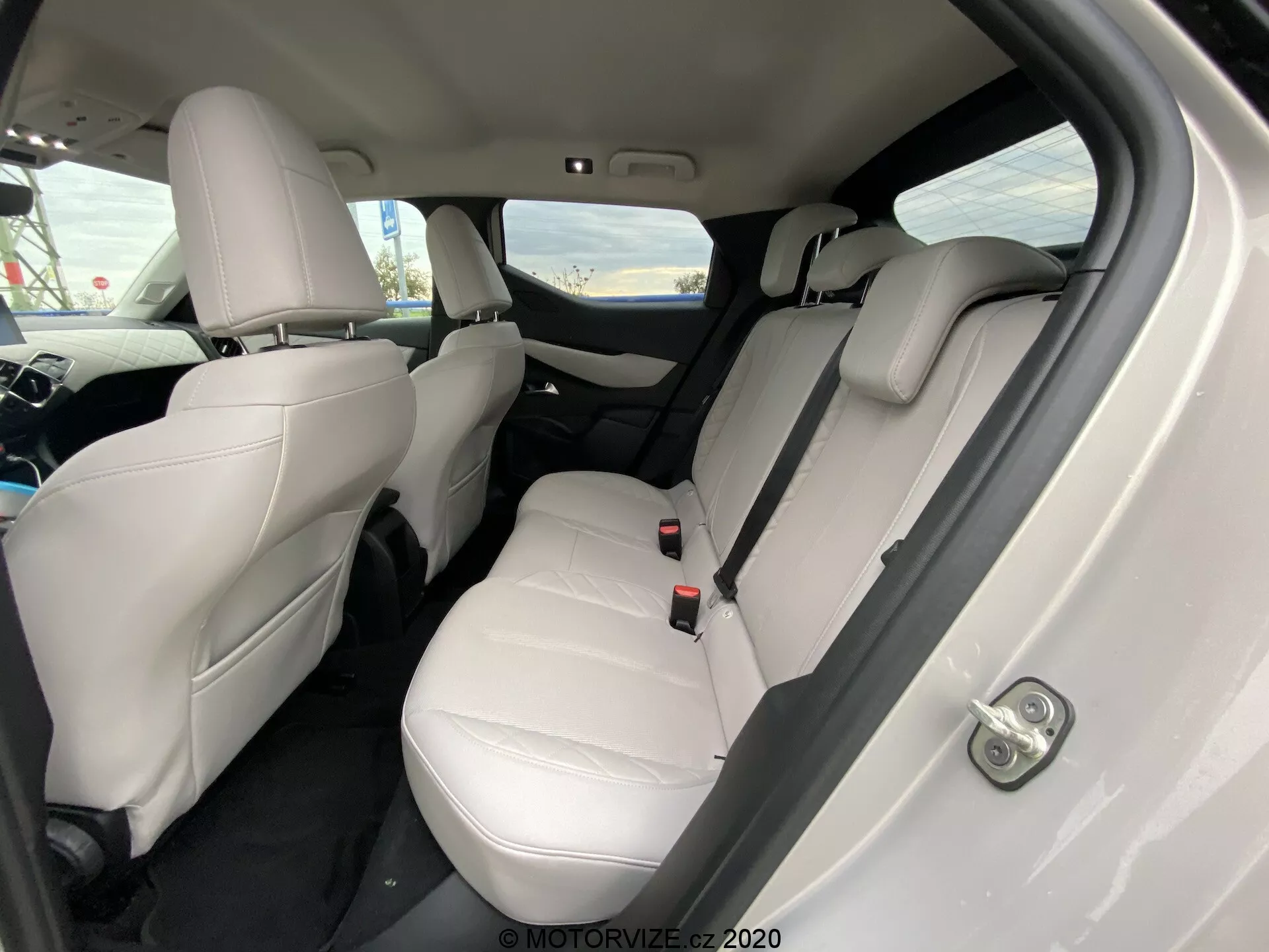 Vue intérieure de la DS Automobiles DS 3 Crossback E-Tense 2019 sous un angle oblique, avec un accent mis sur le compartiment passager arrière. Les sièges en cuir clair, les appuie-tête visibles, les ceintures de sécurité et un accoudoir rabattable sur la banquette arrière signalent un intérieur luxueux et bien entretenu. Les tapis sombres contrastent avec les garnitures claires, et les commandes des fenêtres électriques sont évidentes sur la porte. L'angle de la caméra, légèrement vers le bas, depuis la porte arrière droite, met en valeur l'espace des sièges et le dégagement pour les jambes.