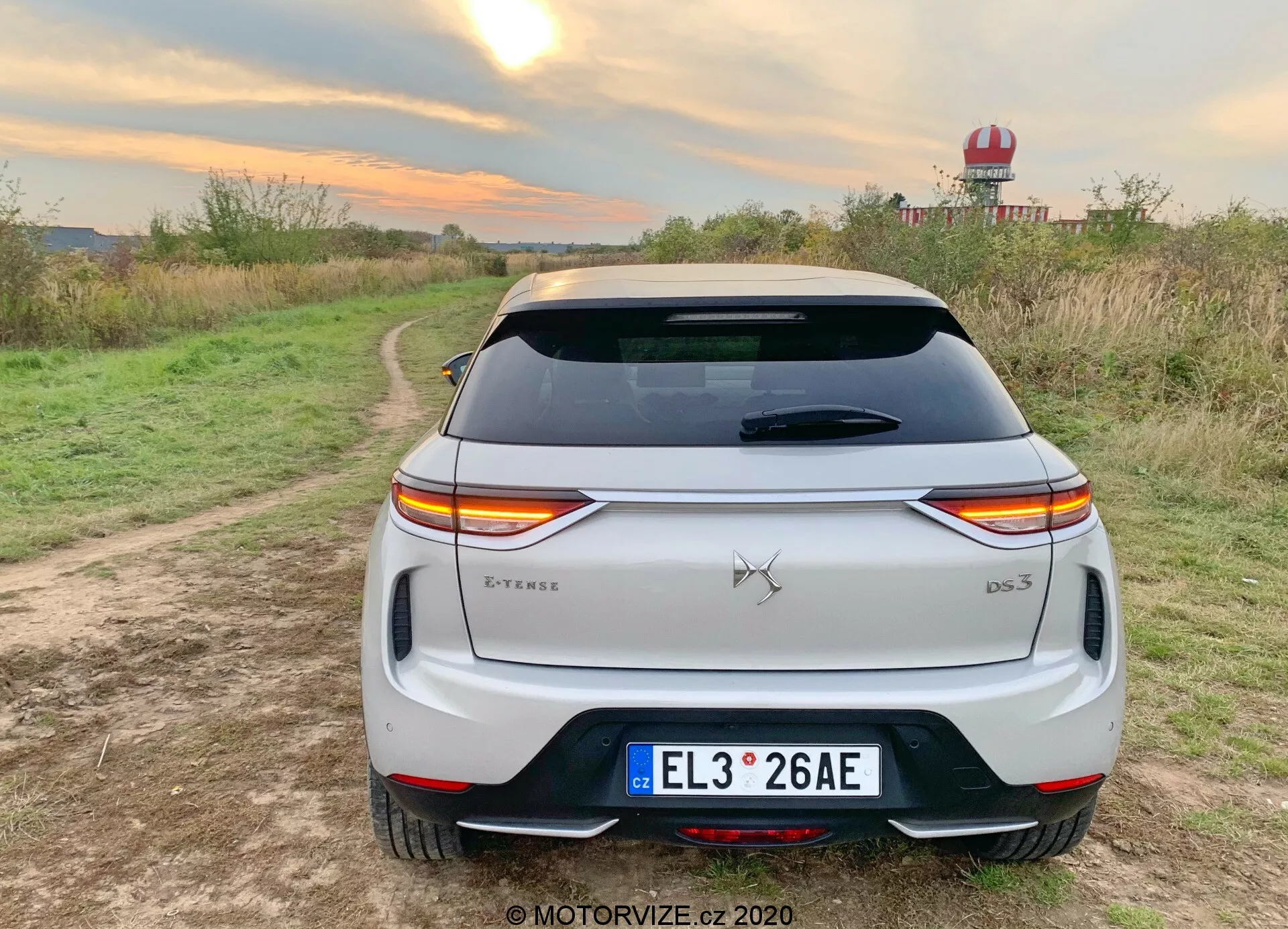 Vue arrière d'un SUV crossover blanc 2019 DS Automobiles DS 3 Crossback E-Tense garé sur un chemin de terre avec de la végétation de chaque côté dans un environnement rural. Les feux arrière LED distinctifs s'étendent sur toute la largeur de l'arrière, interrompus par l'emblème de la marque. Le badge du modèle 'DS 3' est situé en bas à droite du coffre, avec un badge 'E-Tense' en bas à gauche indiquant une motorisation électrique ou hybride. Les caractéristiques incluent une plaque d'immatriculation de style européen, une lunette arrière avec des lignes de dégivrage, un spoiler de toit, un pare-chocs arrière avec des réflecteurs intégrés et une garniture noire, et des doubles sorties d'échappement, suggérant un design plus sportif. L'environnement suggère un essai routier ou une séance photo lifestyle mettant en valeur l'utilisation en extérieur, avec des conditions de lumière chaude mettant en valeur la peinture blanche du véhicule.